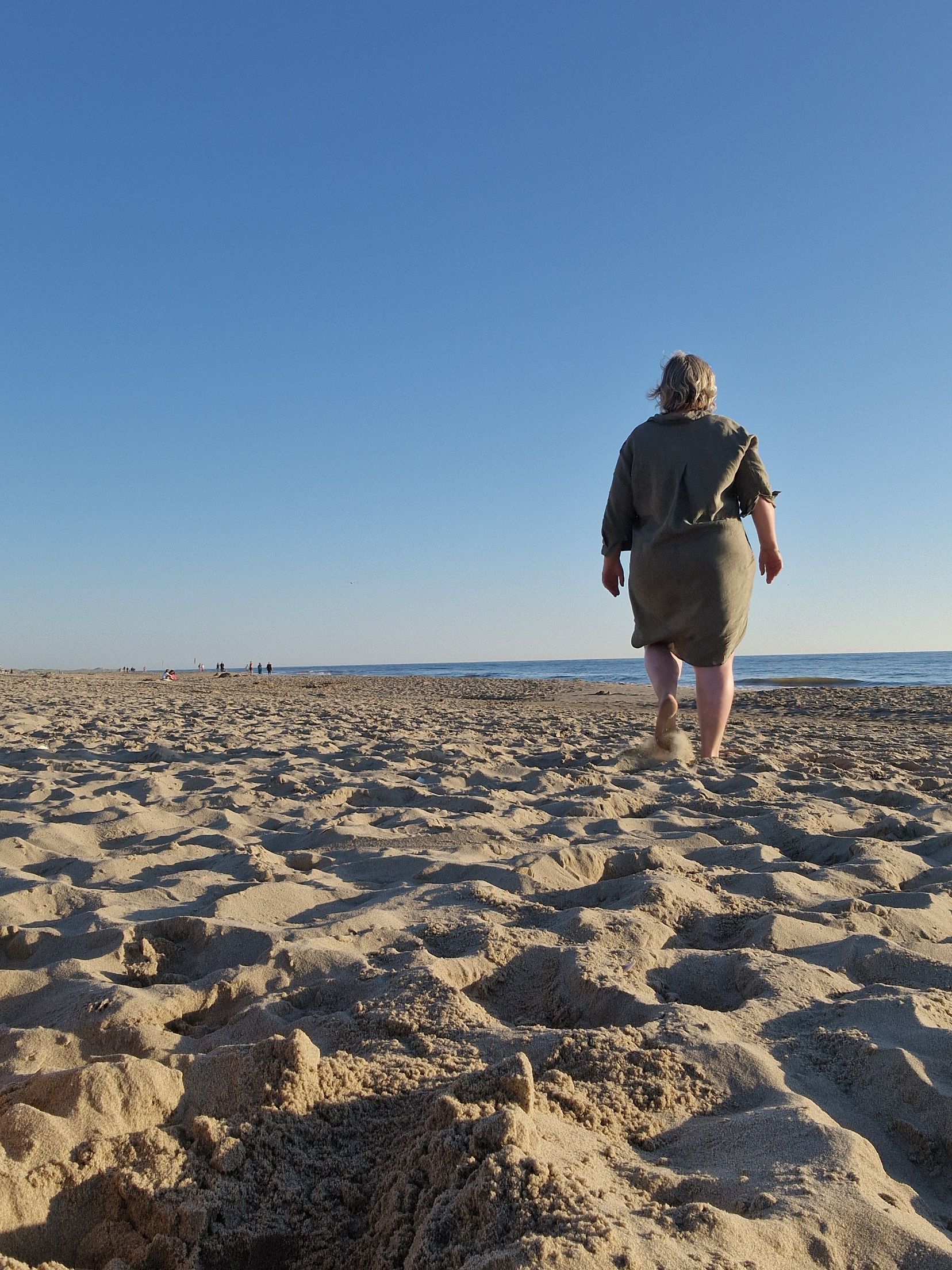 Een persoon loopt over een zandstrand onder een helderblauwe hemel.