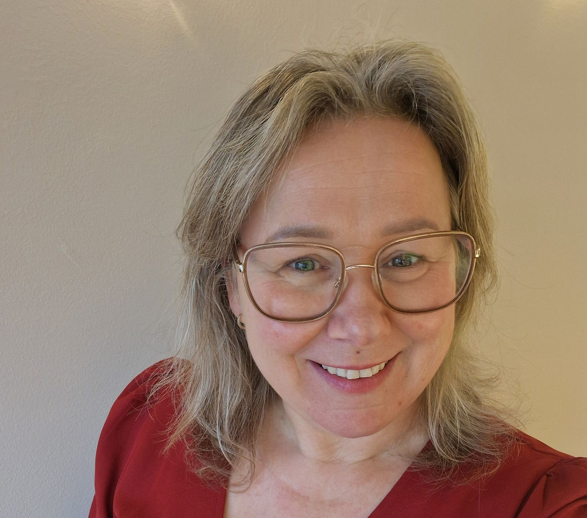Een lachend persoon met een bril en schouderlang haar, gekleed in een rode top, tegen een effen muur.