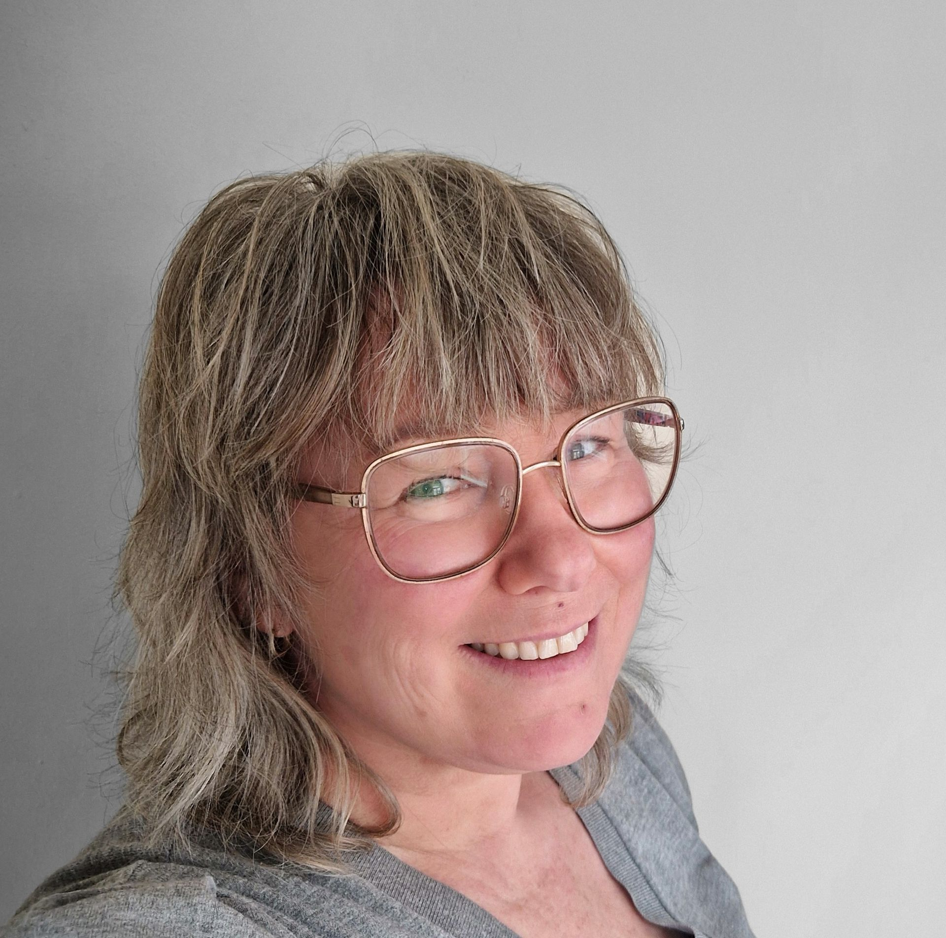 Een lachend persoon met een bril en een grijze trui tegen een effen muur.