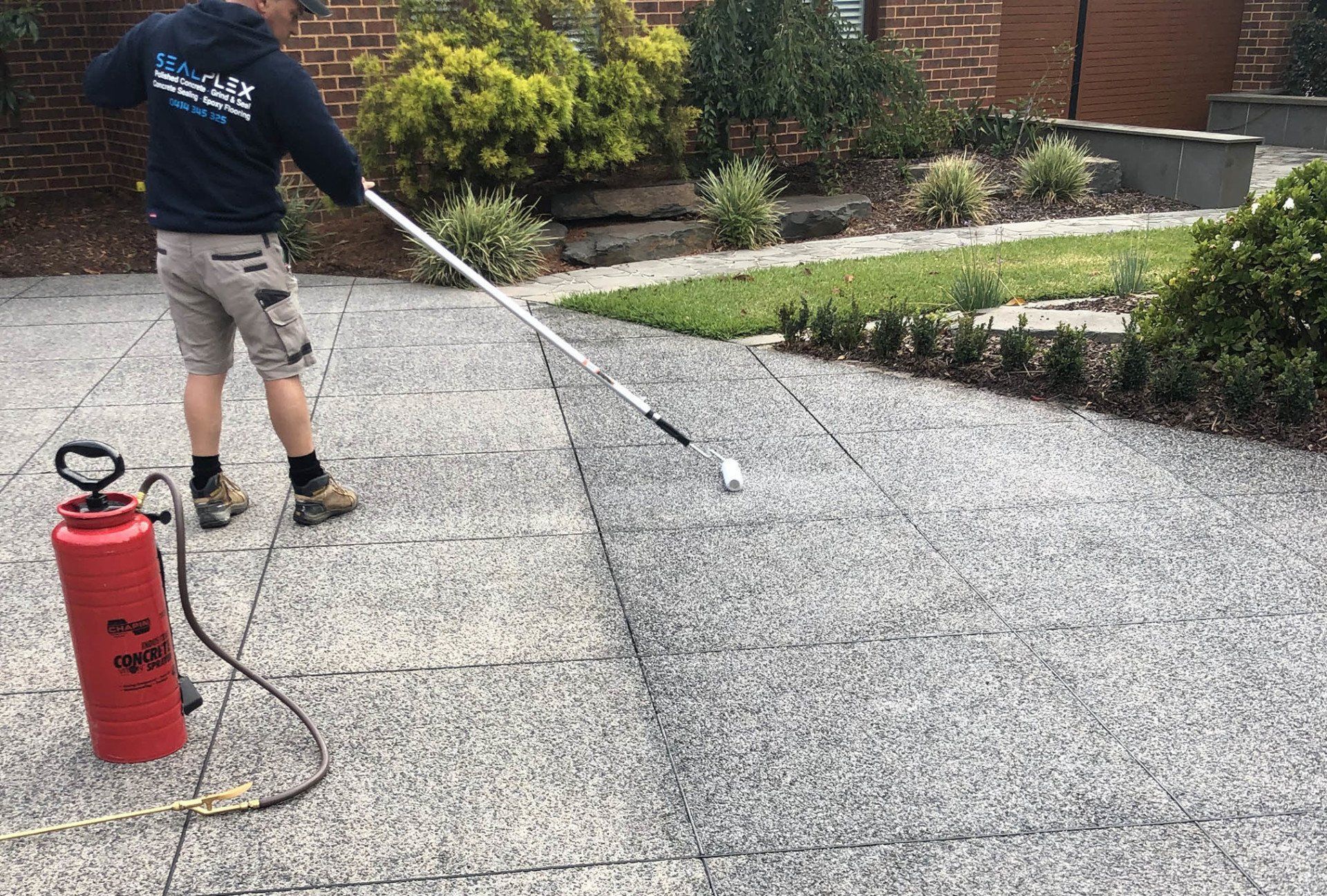 Man spraying a concrete driveway with a red sprayer; exterior setting with greenery and a house.