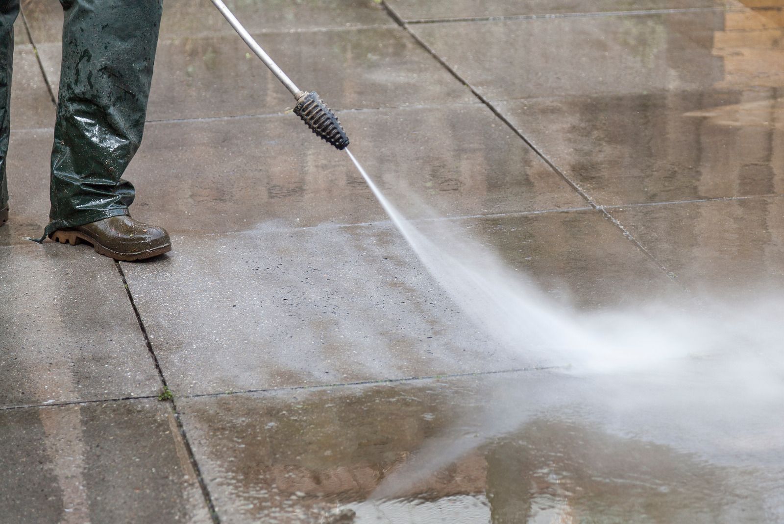 Person pressure-washing a concrete sidewalk, with water spraying across the wet pavement.