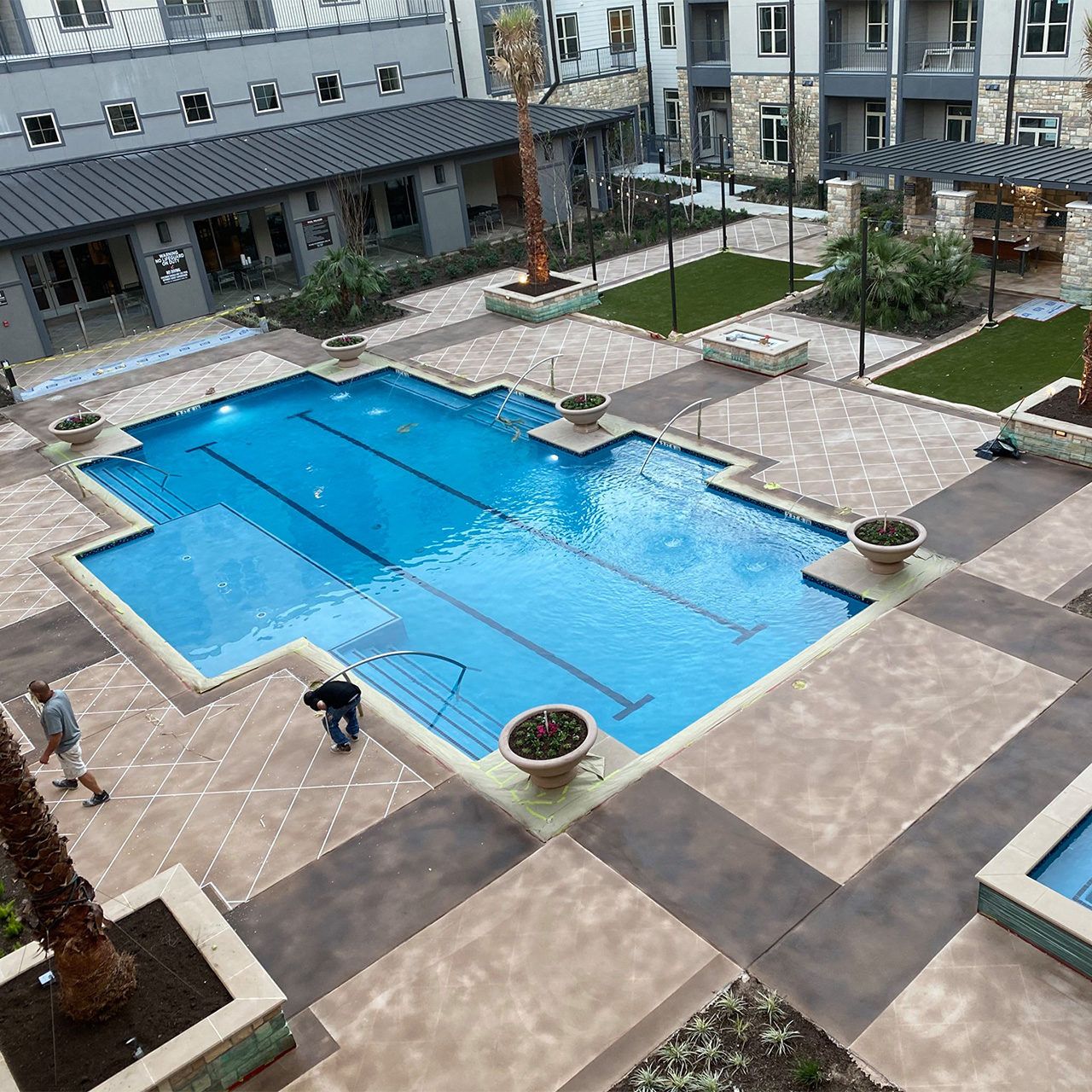 Outdoor courtyard with a blue swimming pool, beige stone deck, and a few people walking nearby.