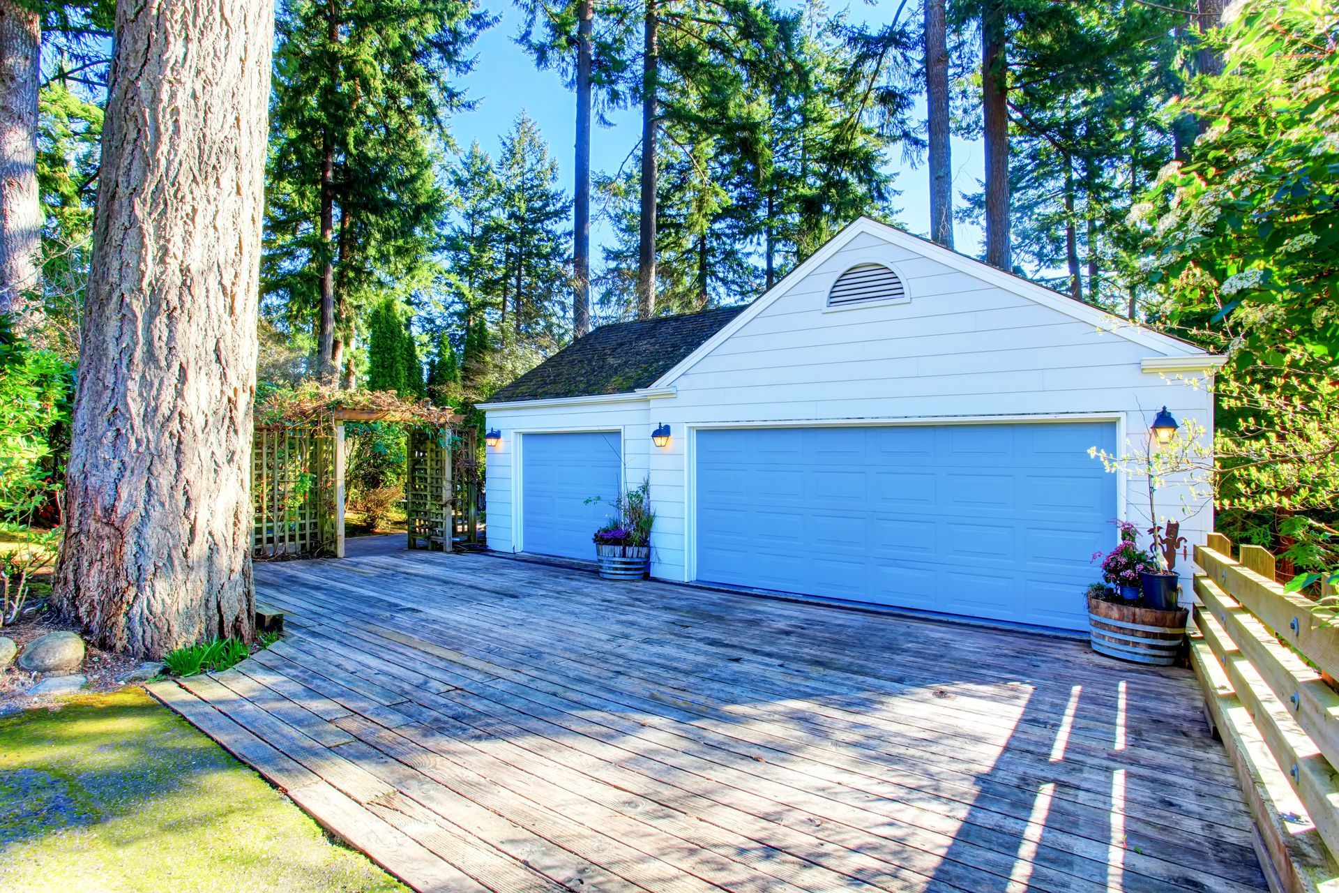 White garage with blue doors beside a large tree on a sunny wooded driveway
