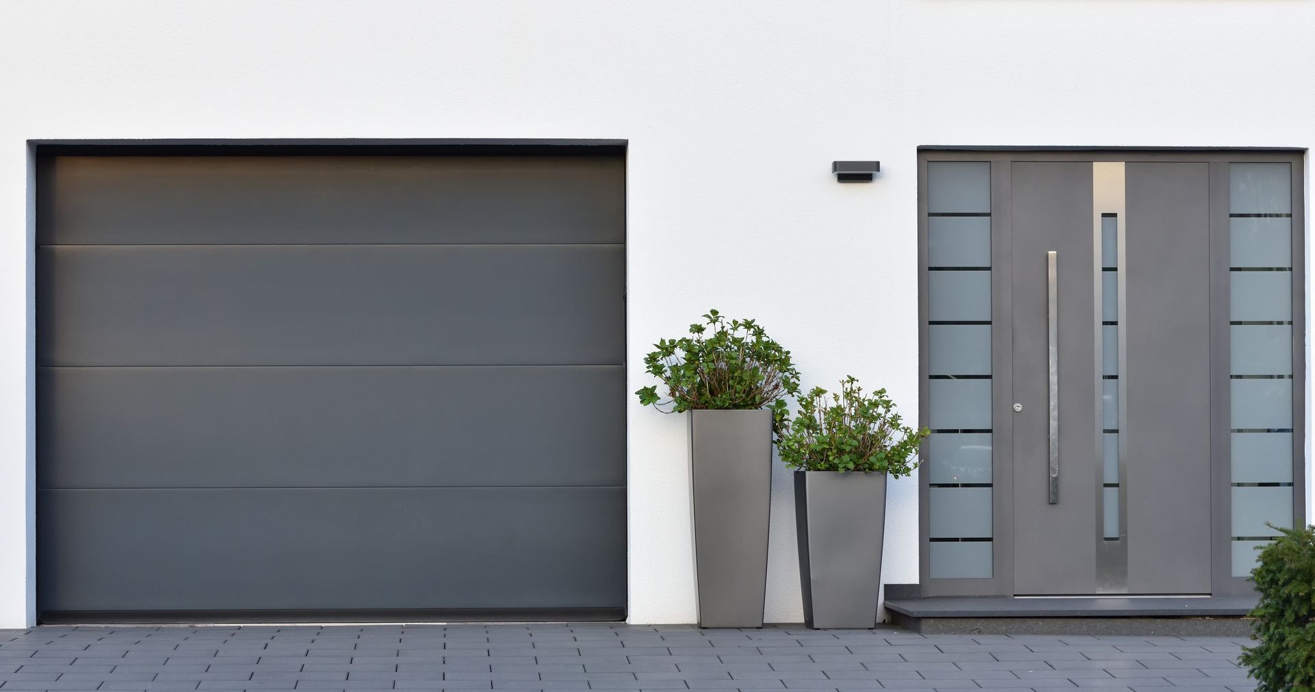 Modern gray house facade with a dark garage door, front entrance, and potted plants by the door
