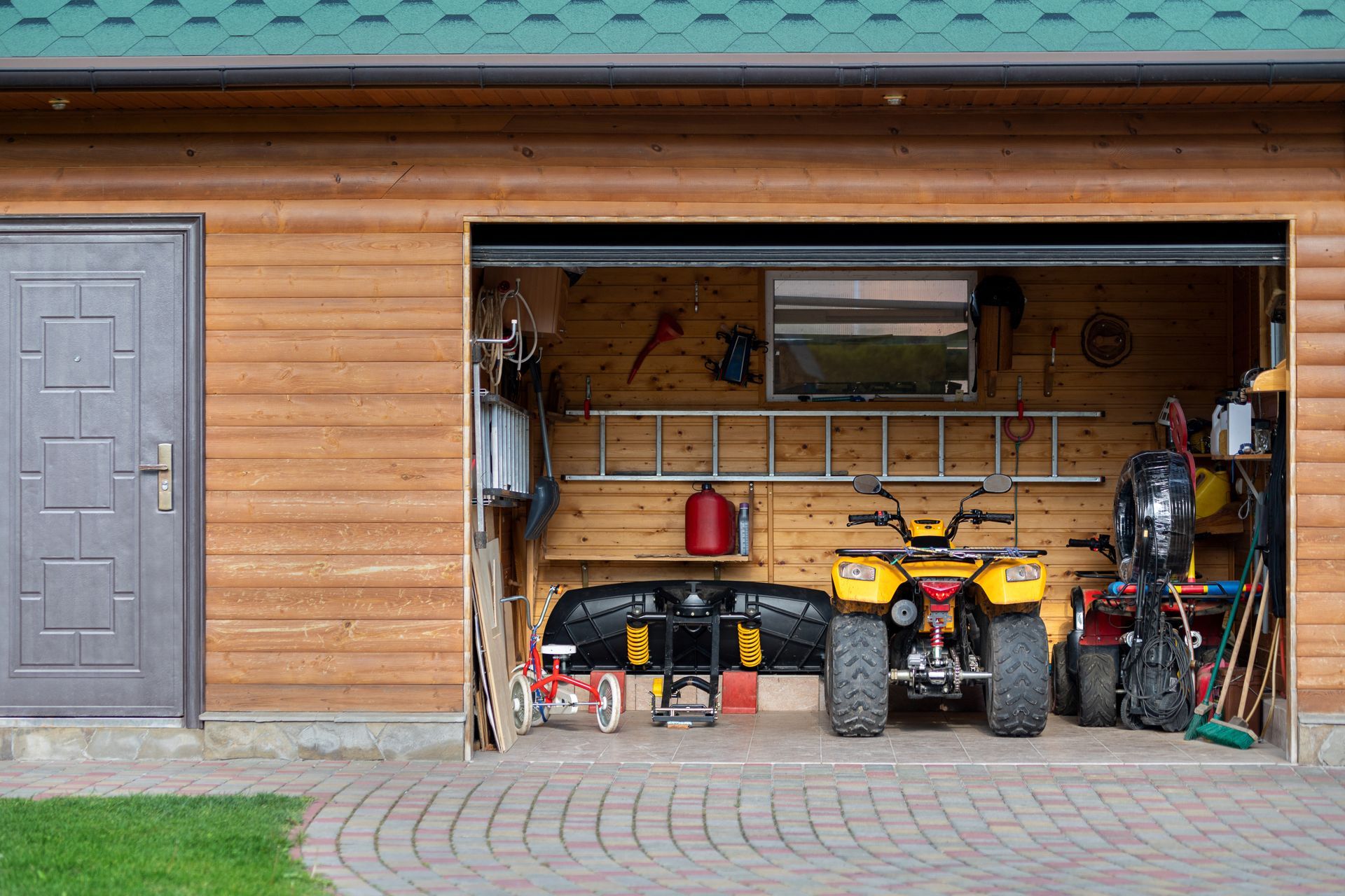 Wooden garage with an ATV, lawn mower, tools, and bikes inside beside a gray door