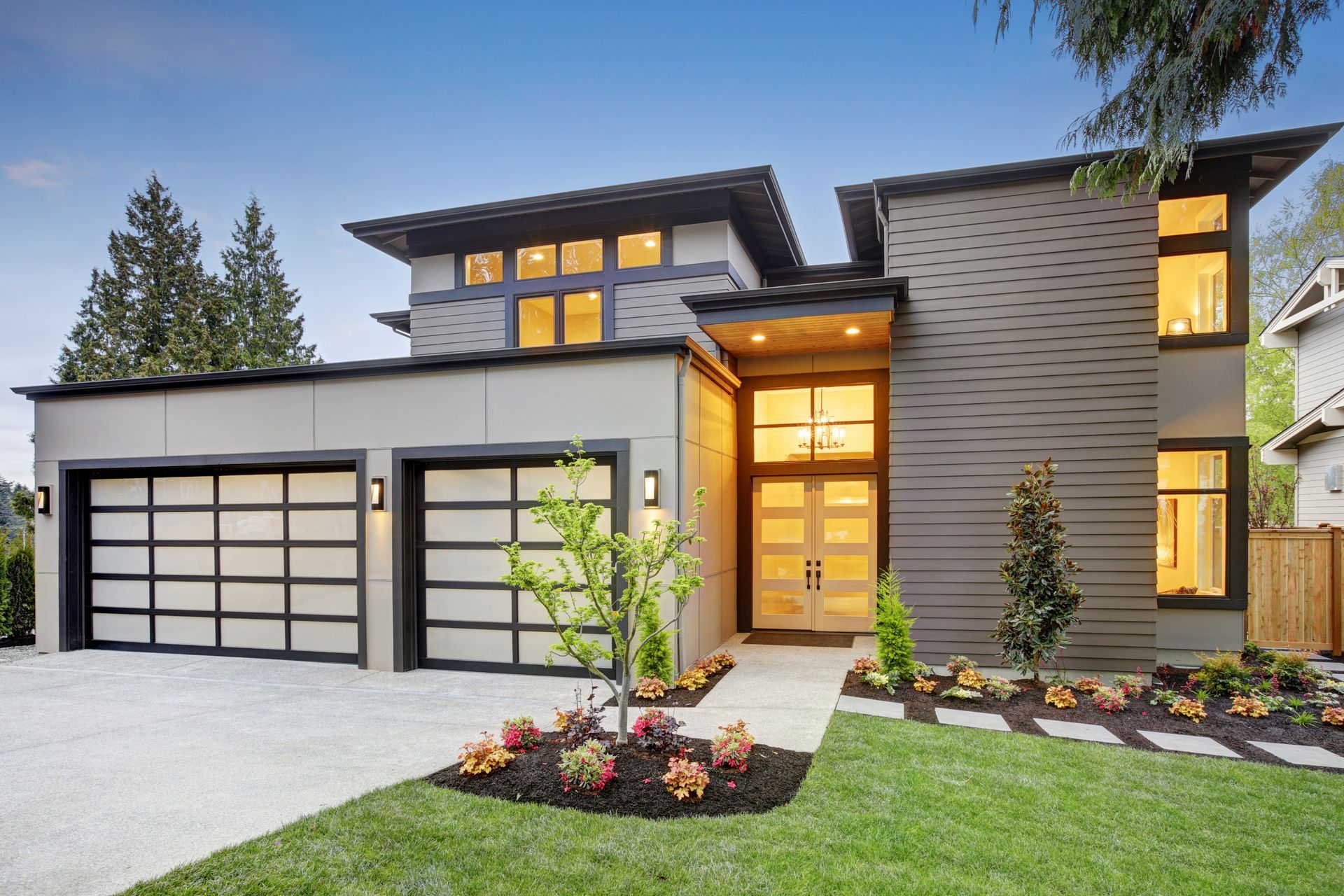 Modern two-story house with a white garage, warm entry lights, and landscaped front yard at dusk