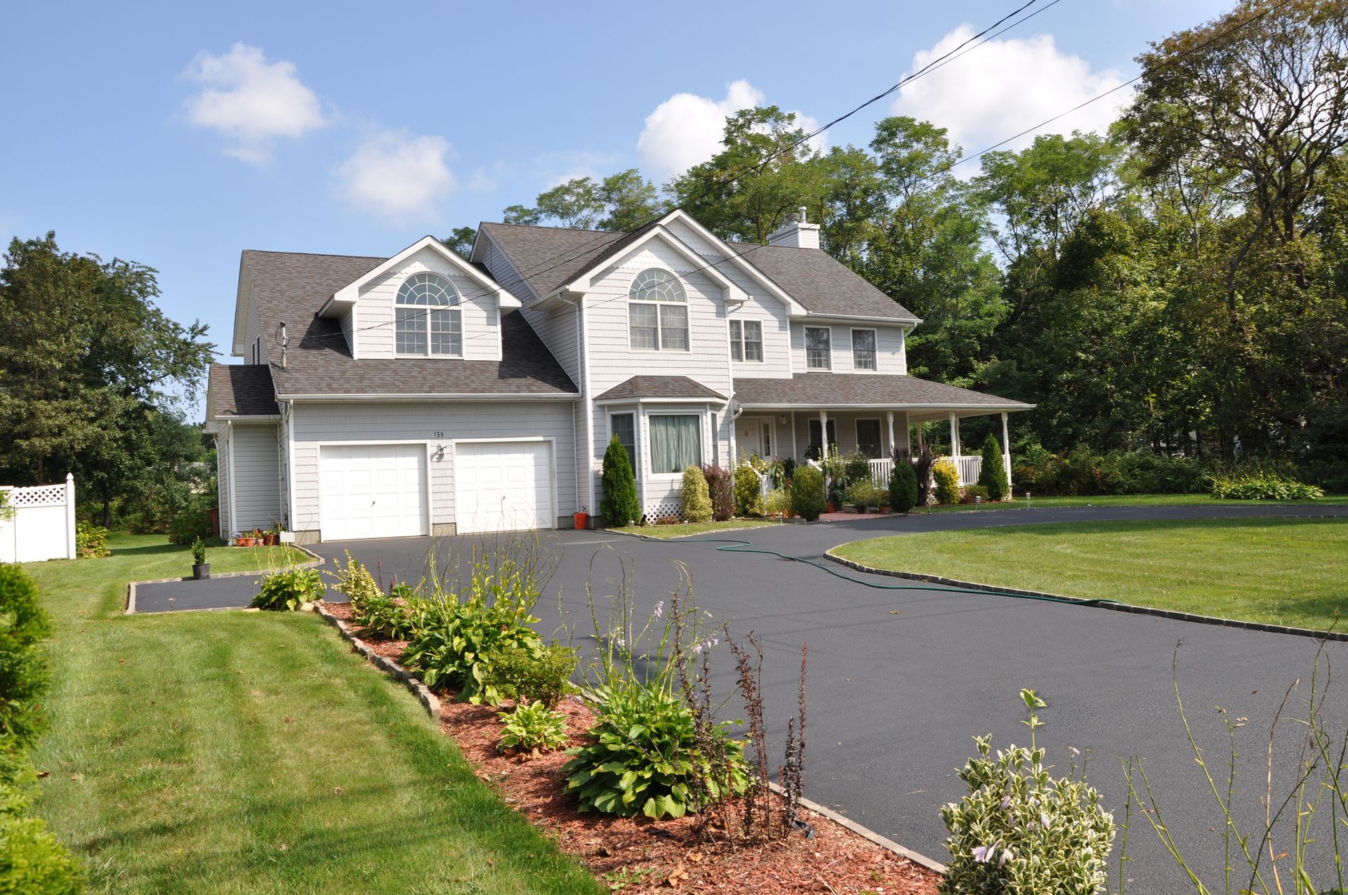Suburban two-story gray house with attached garage, landscaped lawn, and long blacktop driveway under clear sky
