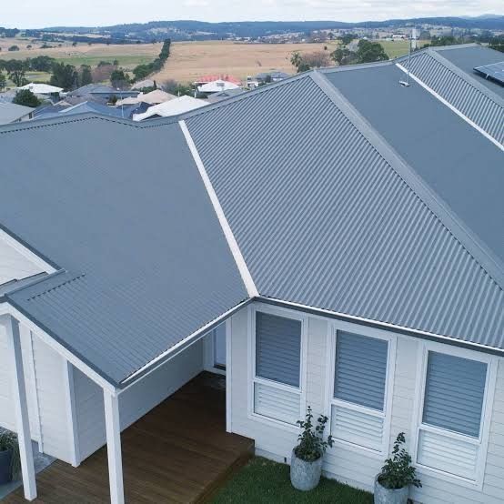 Light grey house with a large corrugated metal roof in a suburban neighborhood, featuring clean white trim and views of distant rolling fields. — Apex Roofing Group in Maclean, NSW