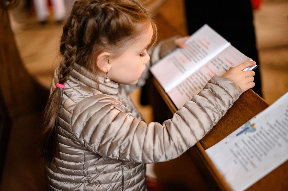 Child in a beige quilted jacket reading a book at a wooden lectern indoors