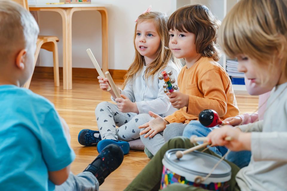 Children sitting on a wooden floor, playing instruments in a bright classroom circle time.