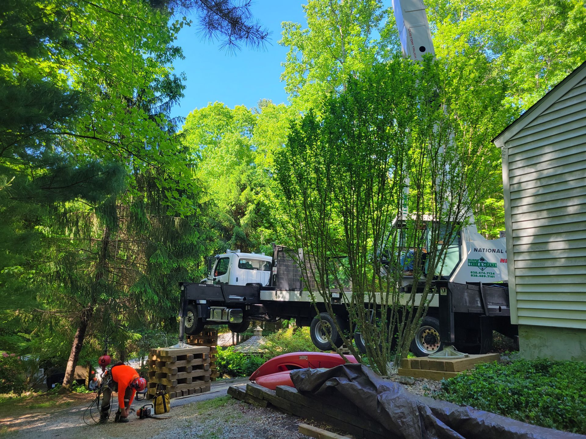 Truck with crane lifting pallets in a wooded yard with worker nearby; green trees, blue sky.