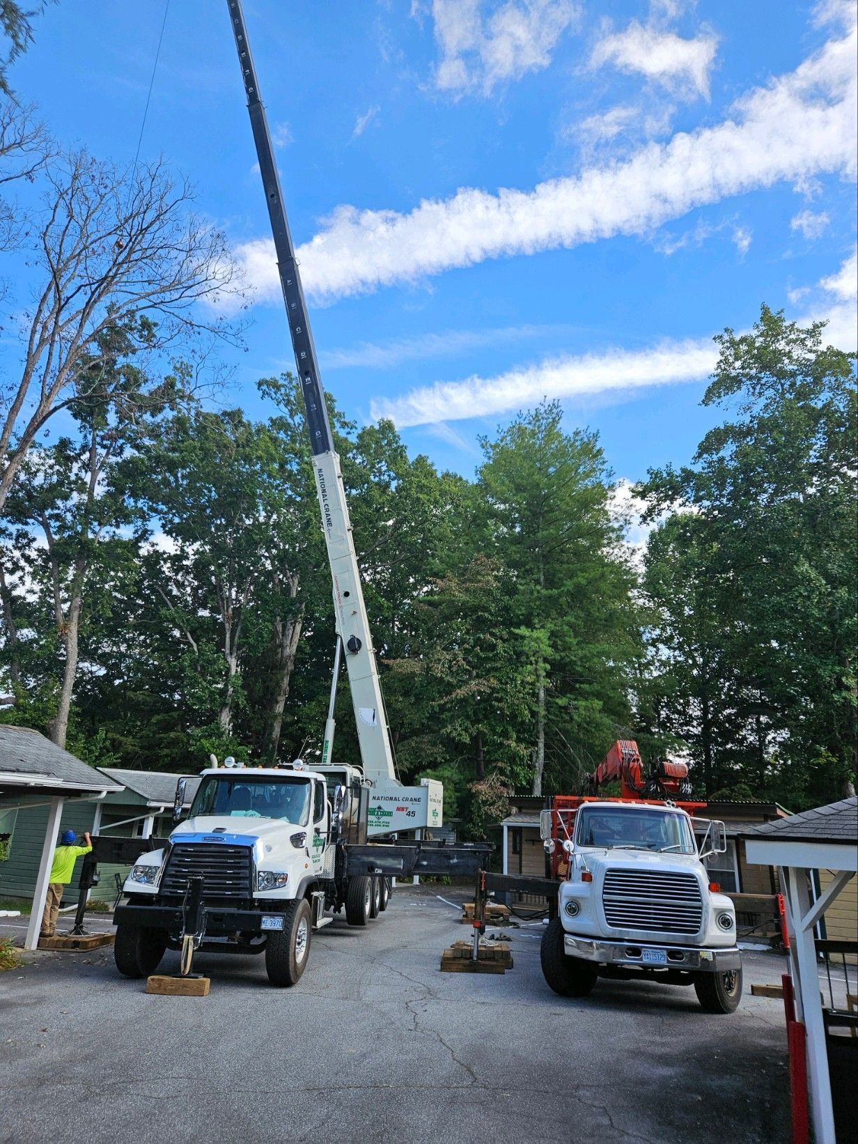 Crane truck and other truck parked near trees under a blue sky, possibly construction site.