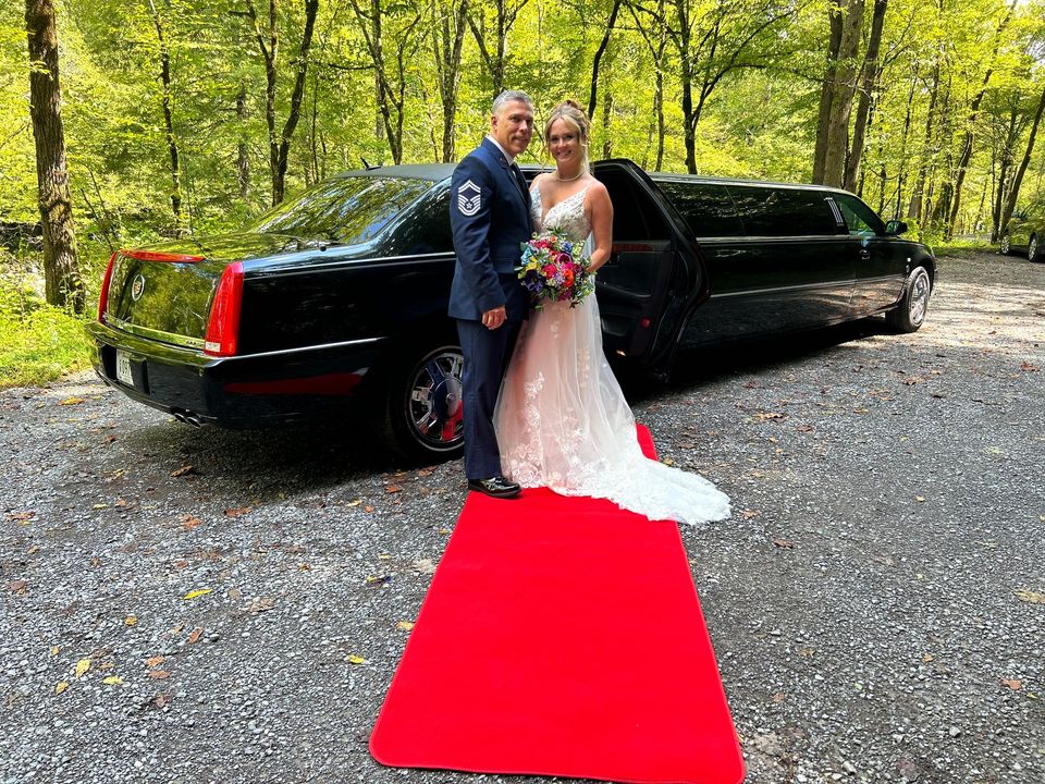 Couple in wedding attire posing beside a black limousine on a red carpet in a wooded outdoor setting