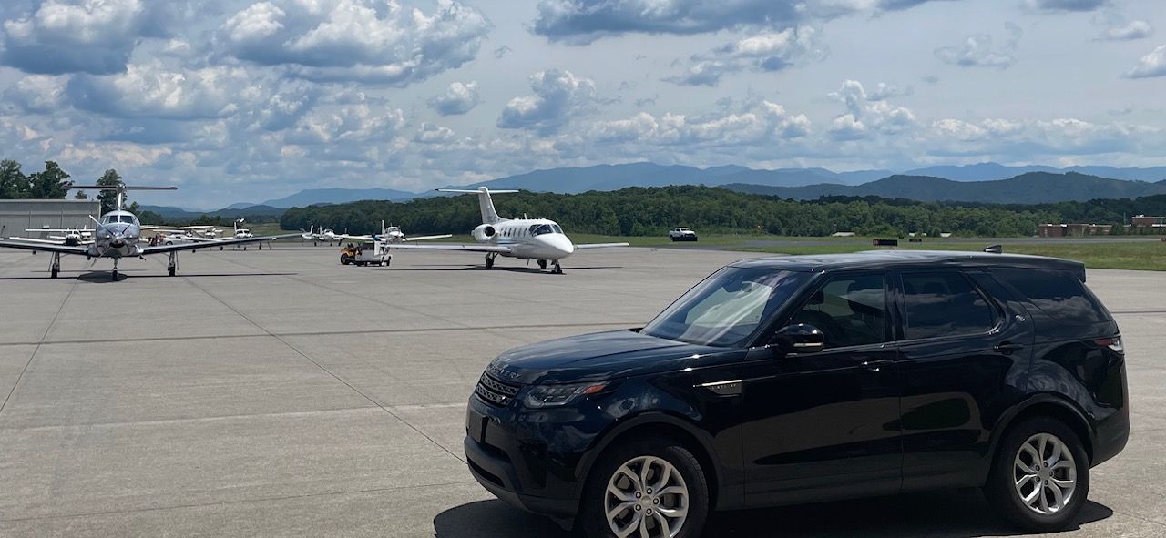 Black SUV parked on a runway with several small planes and distant mountains under a cloudy sky.