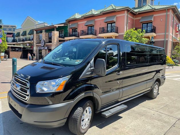 Black Ford passenger van parked in front of a brick building on a sunny street.