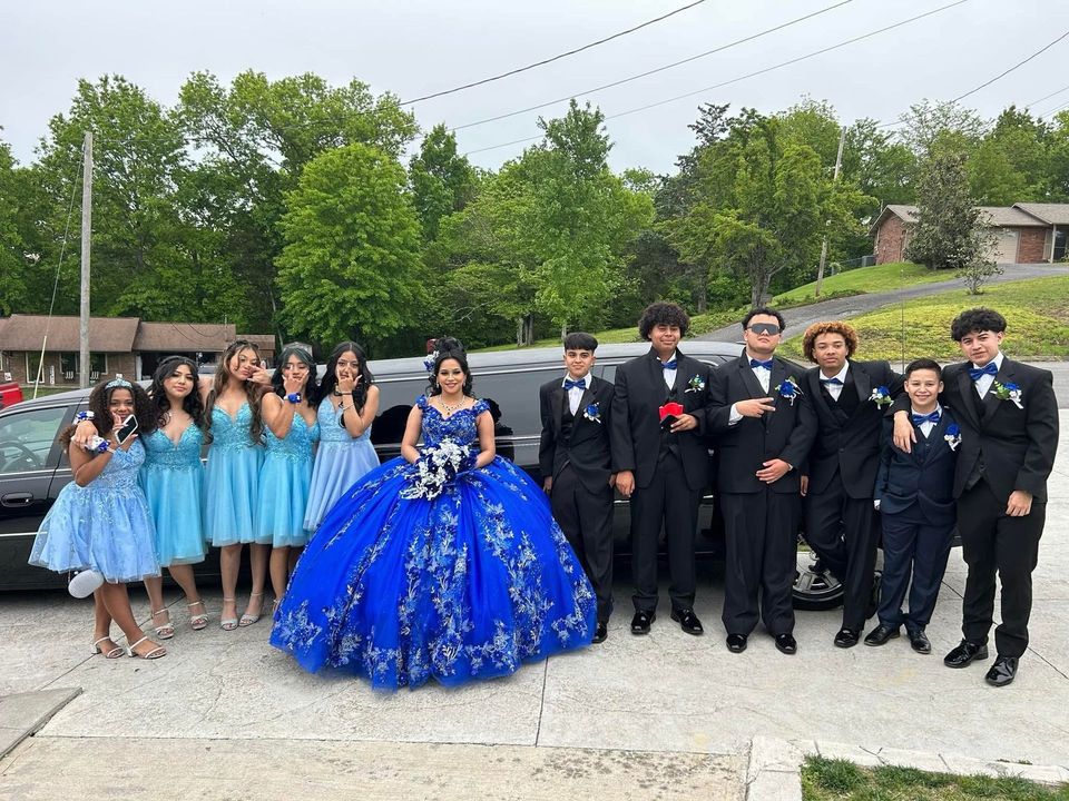 Group in blue gowns and black suits posing outdoors with a royal blue ball gown in front