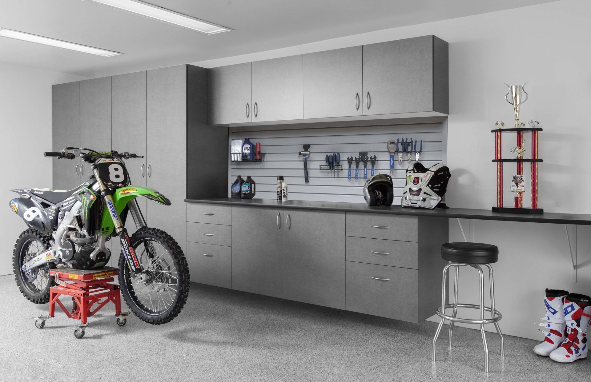 Garage workshop with a green dirt bike on a lift, cabinets, workbench, and stools.