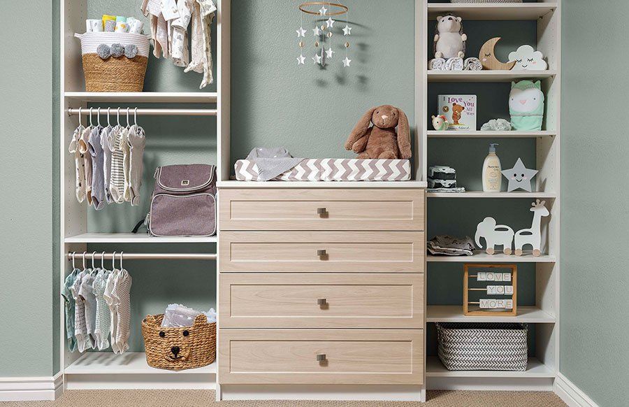 Nursery storage wall with pale wood drawers, shelves, and baby clothes in soft mint and cream tones