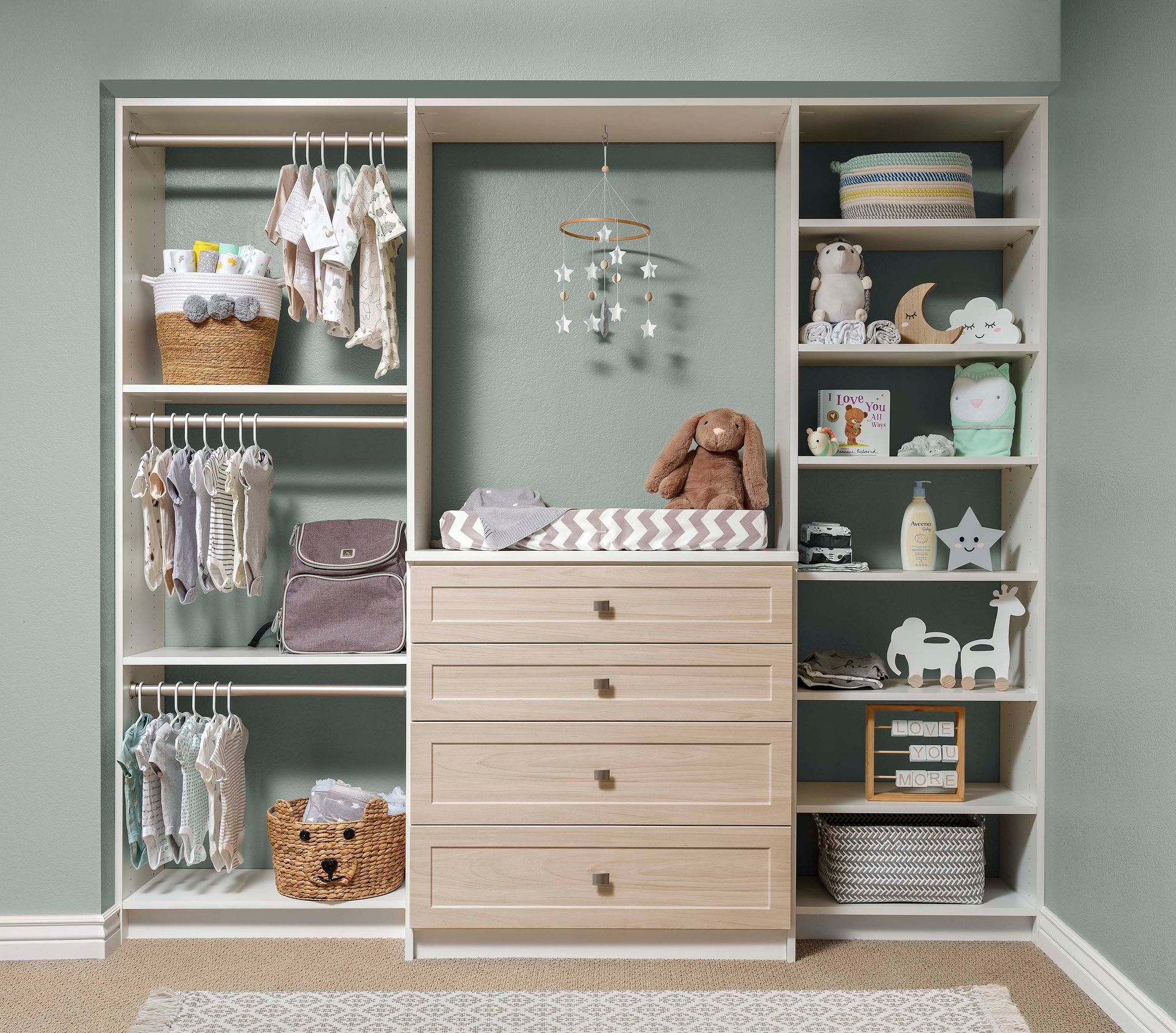 Built-in nursery closet with light wood drawers, shelves, baskets, clothes, and white decor on sage walls