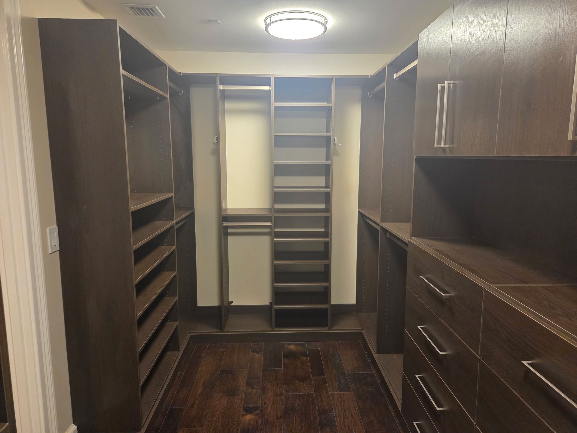Walk-in closet with dark wood shelving, drawers, and hanging space, lit by a ceiling light.