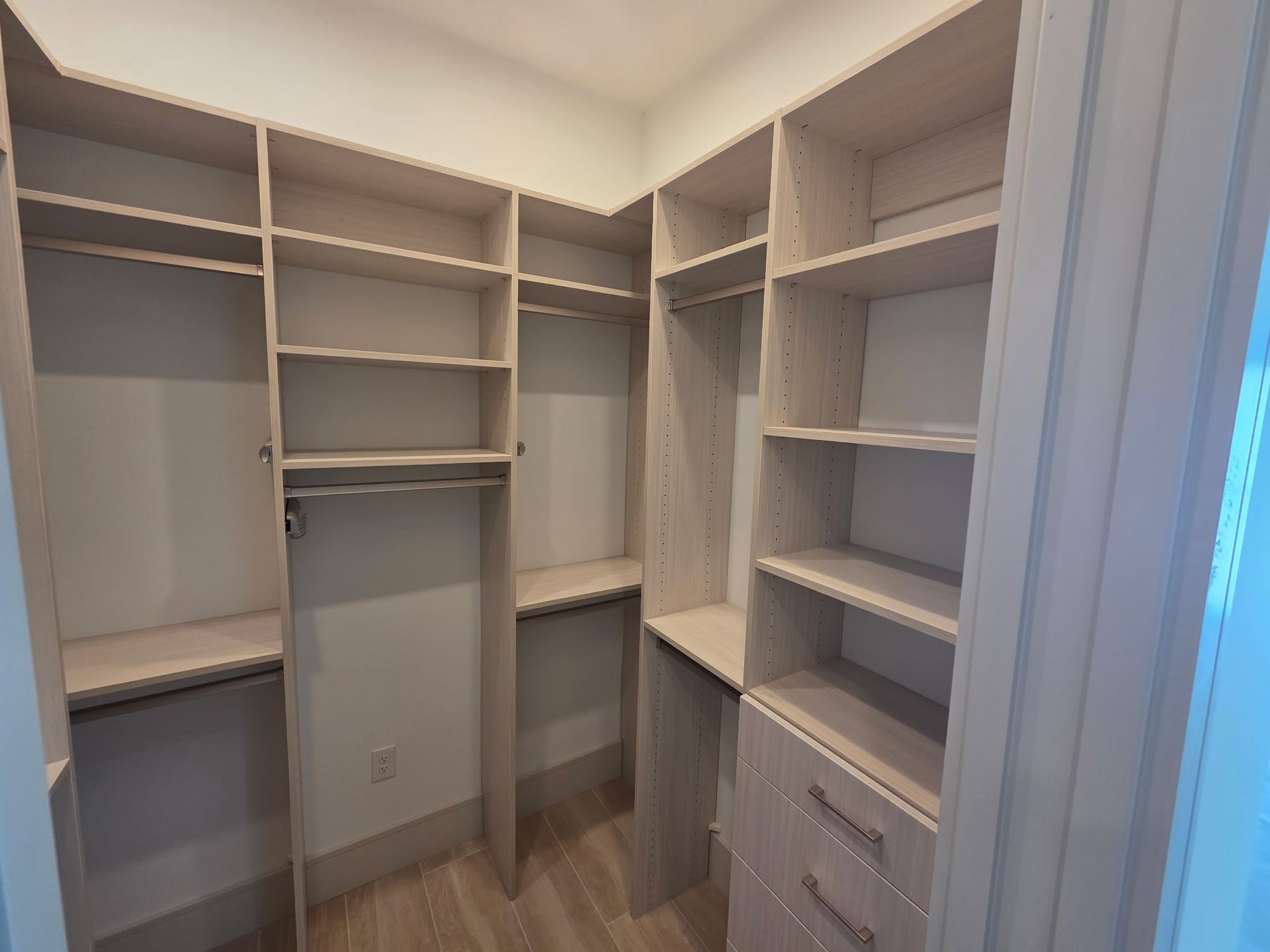 Empty walk-in closet with white built-in shelves, hanging rods, and drawers in a beige room