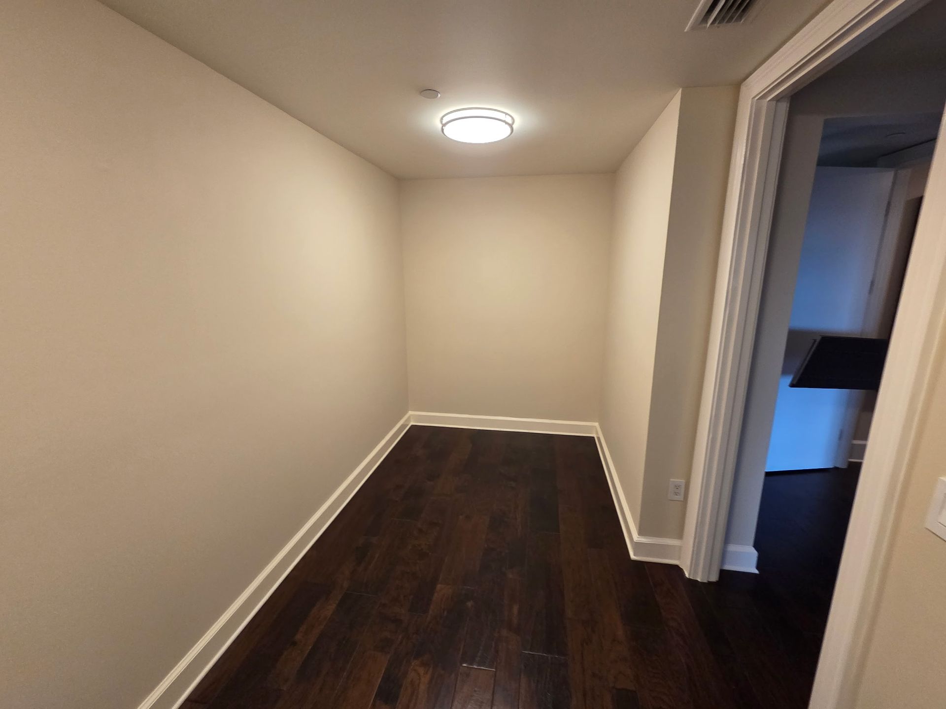 Narrow hallway with beige walls, dark wood floor, and a doorway to a blue-lit room.