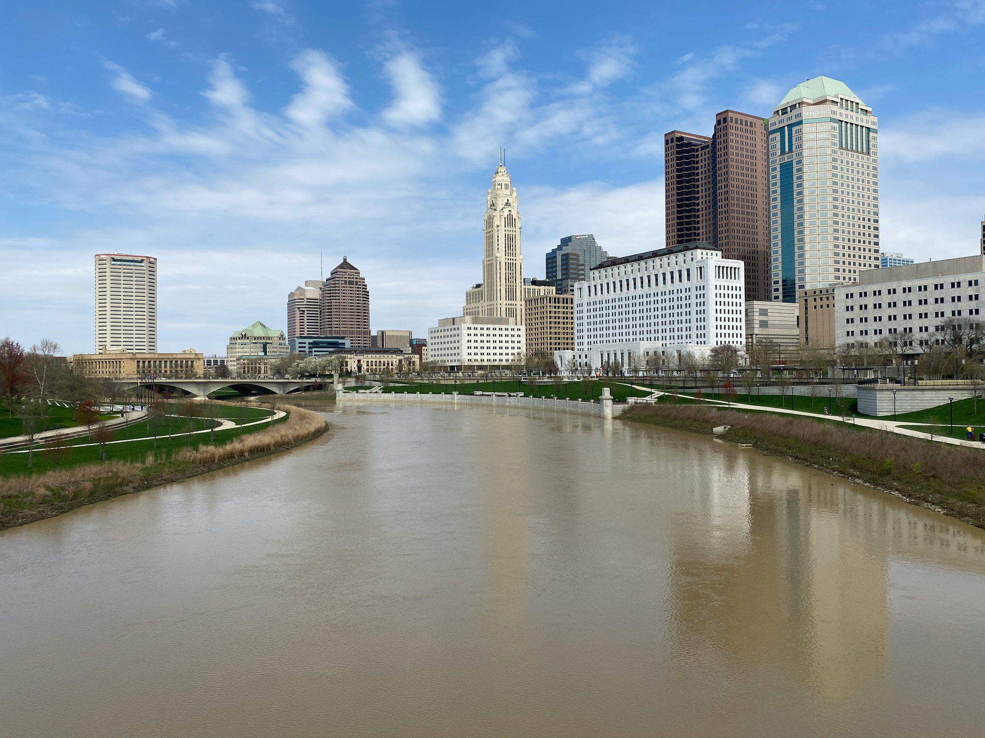 Riverfront city skyline under a blue sky, with tall buildings along a calm brown river.