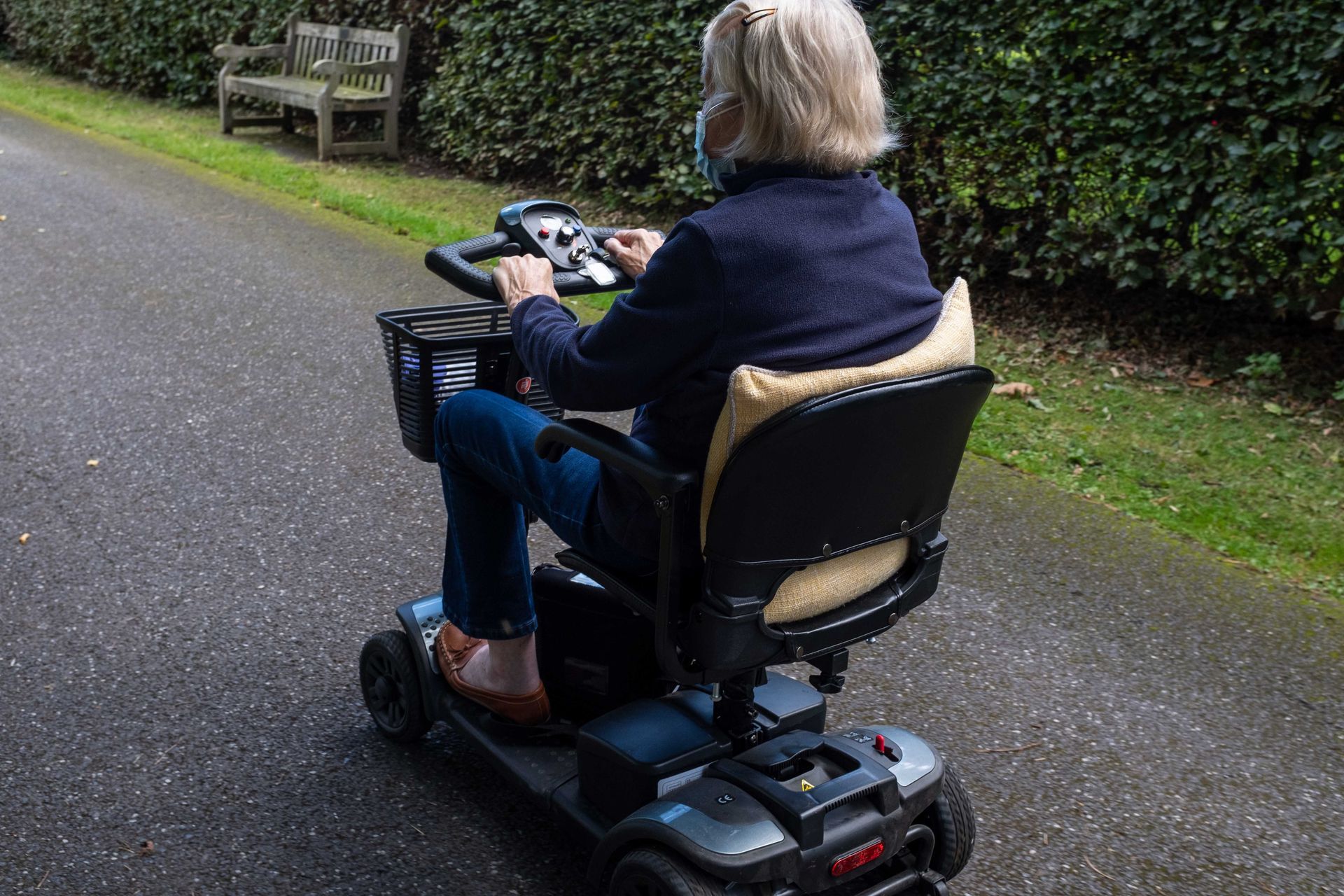 Person driving a mobility scooter on a paved path beside a hedge and bench Person driving a mobility scooter on a paved path beside a hedge and bench