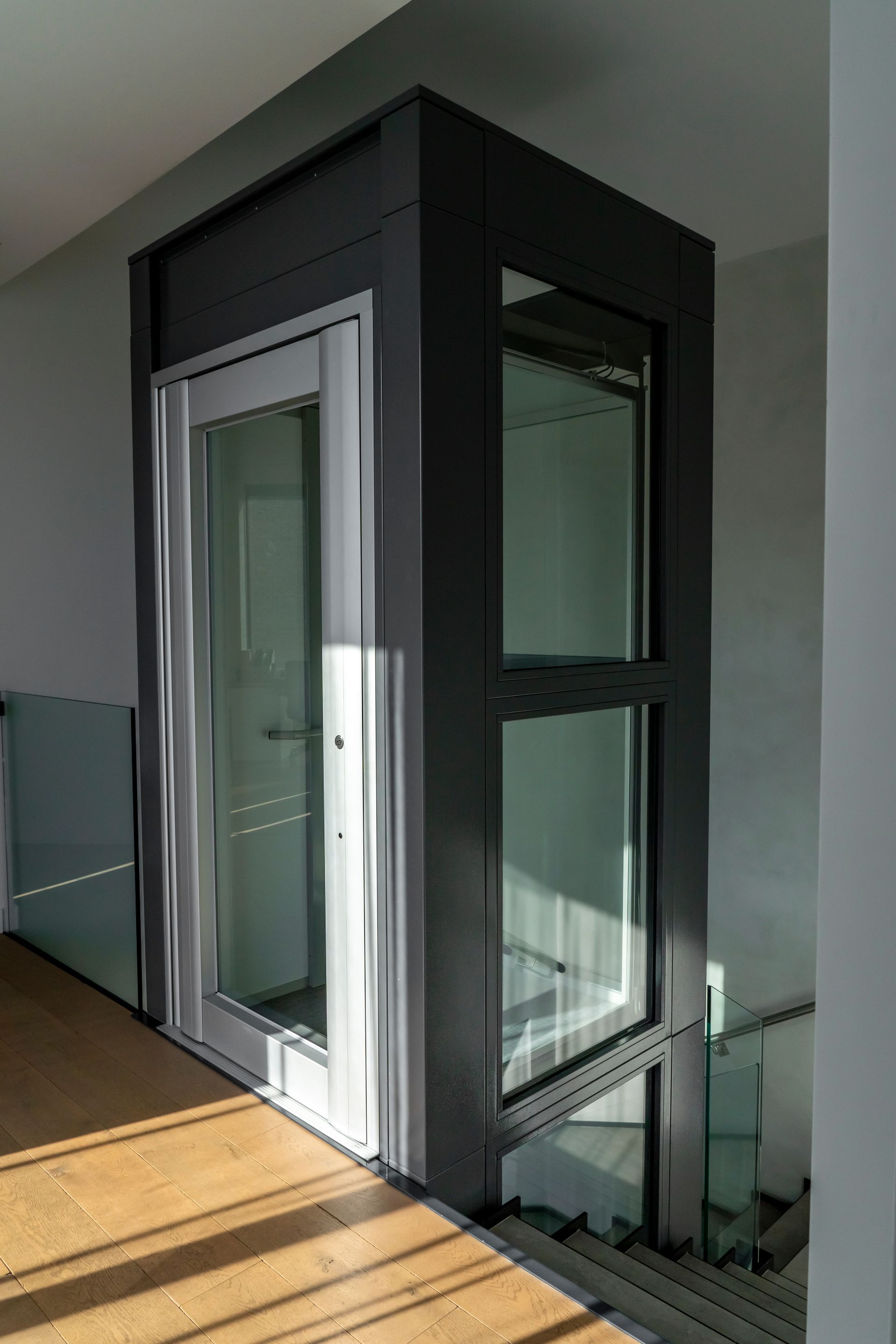 Glass and black-frame home elevator in a bright hallway with wood flooring.