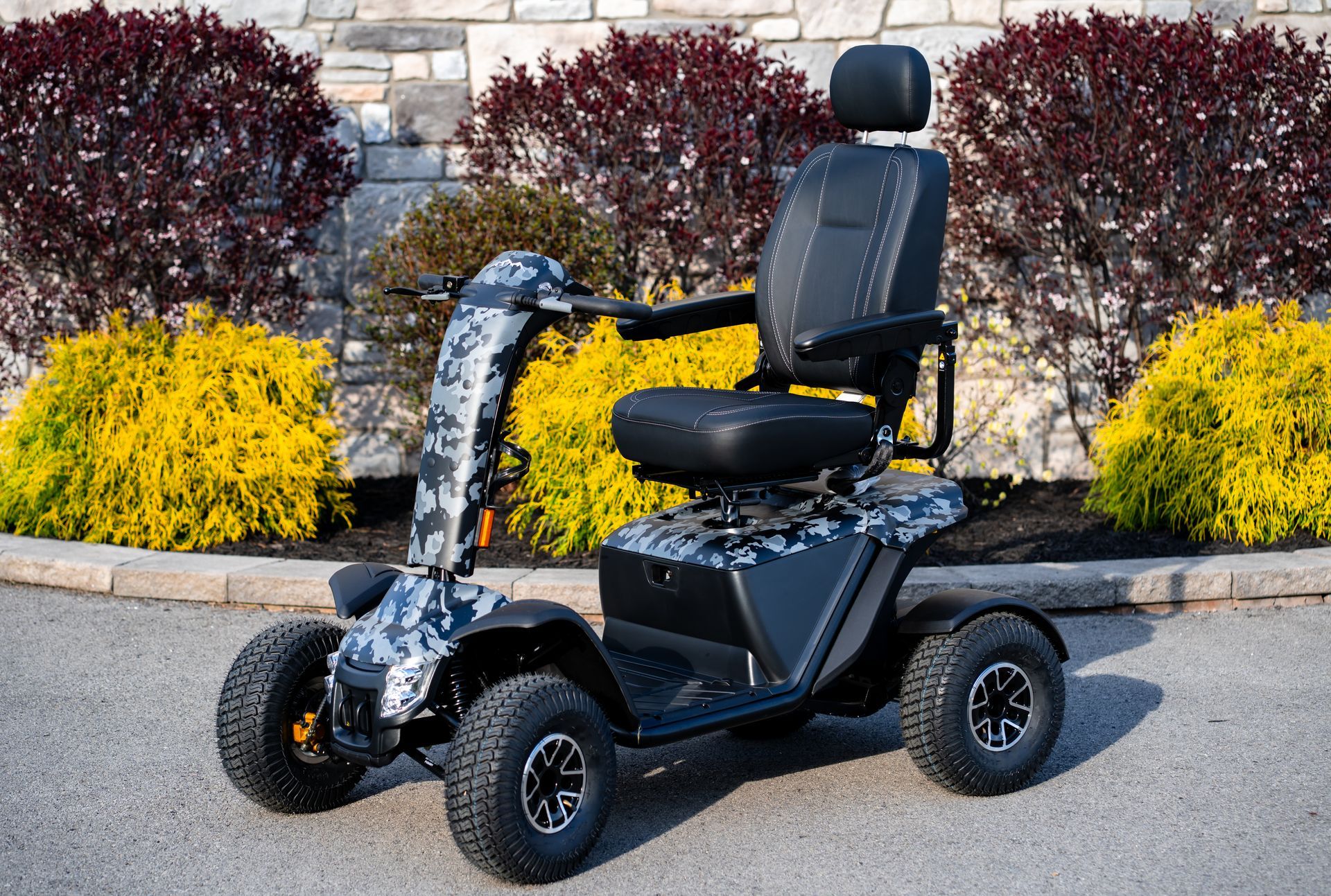 Off-road mobility scooter with camo accents and black seat. Outdoors, in front of bushes.
