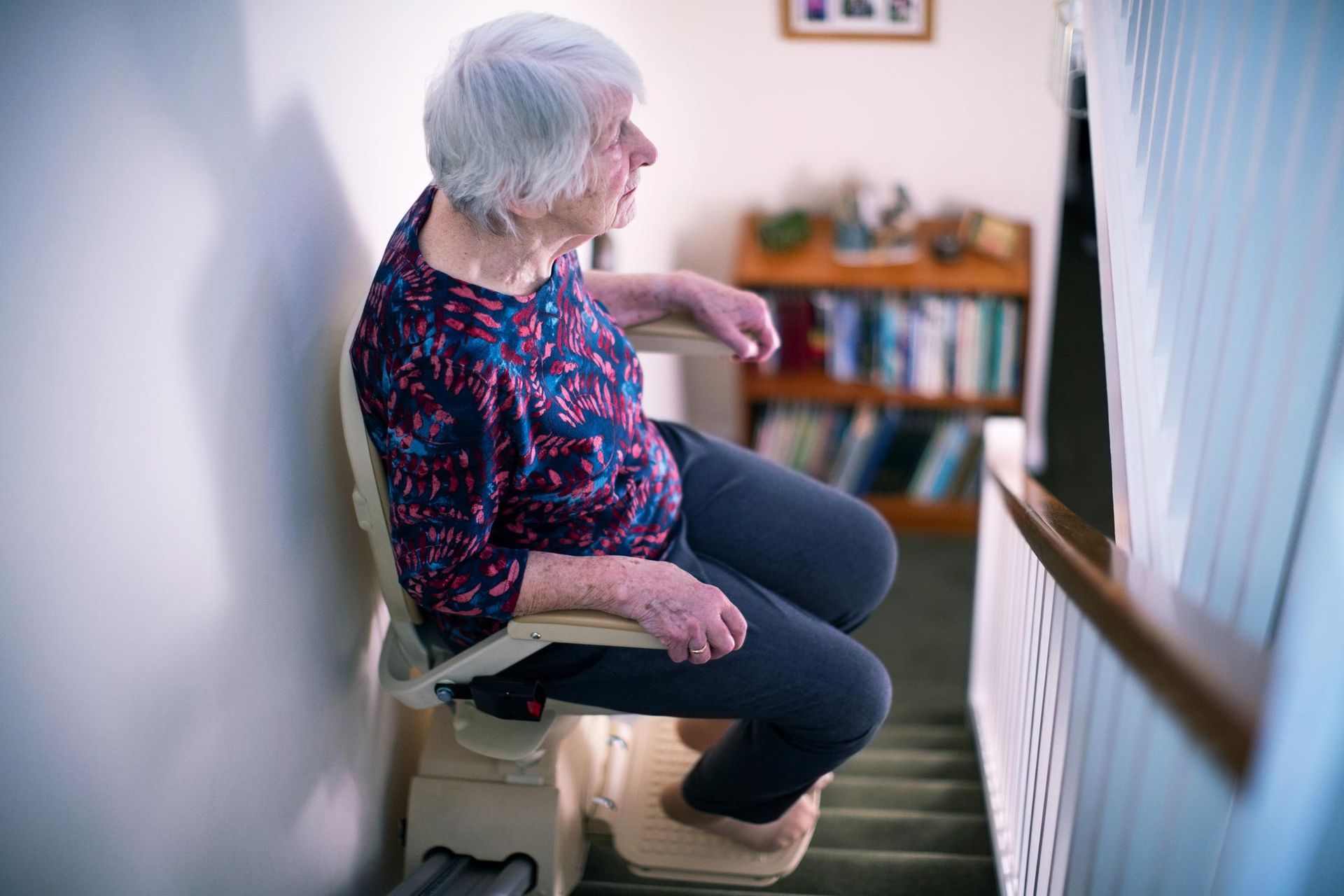 Elderly woman riding stairlift chair up residential home staircase for mobility.