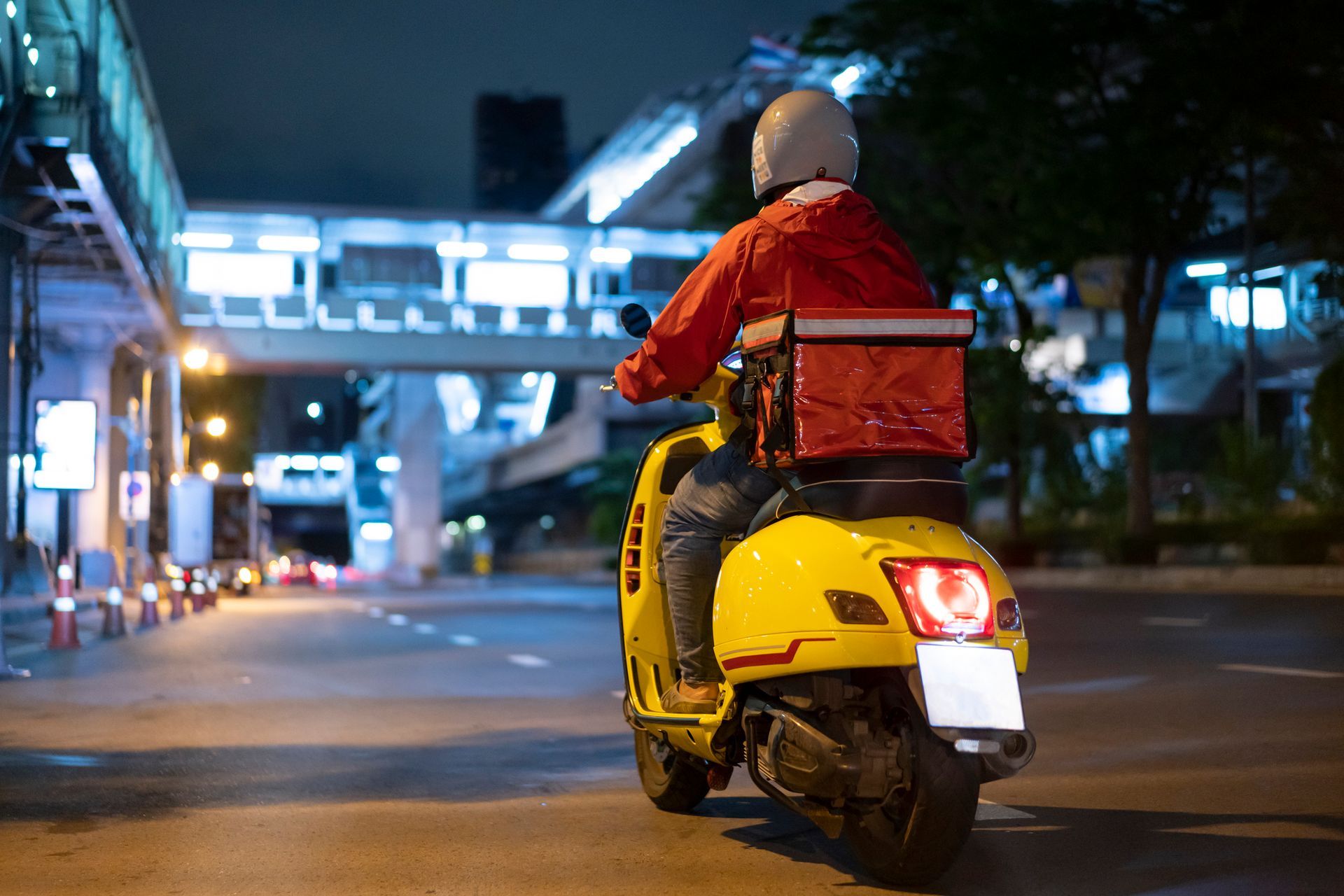 A person on a yellow scooter delivering food at night, wearing a red jacket and helmet.