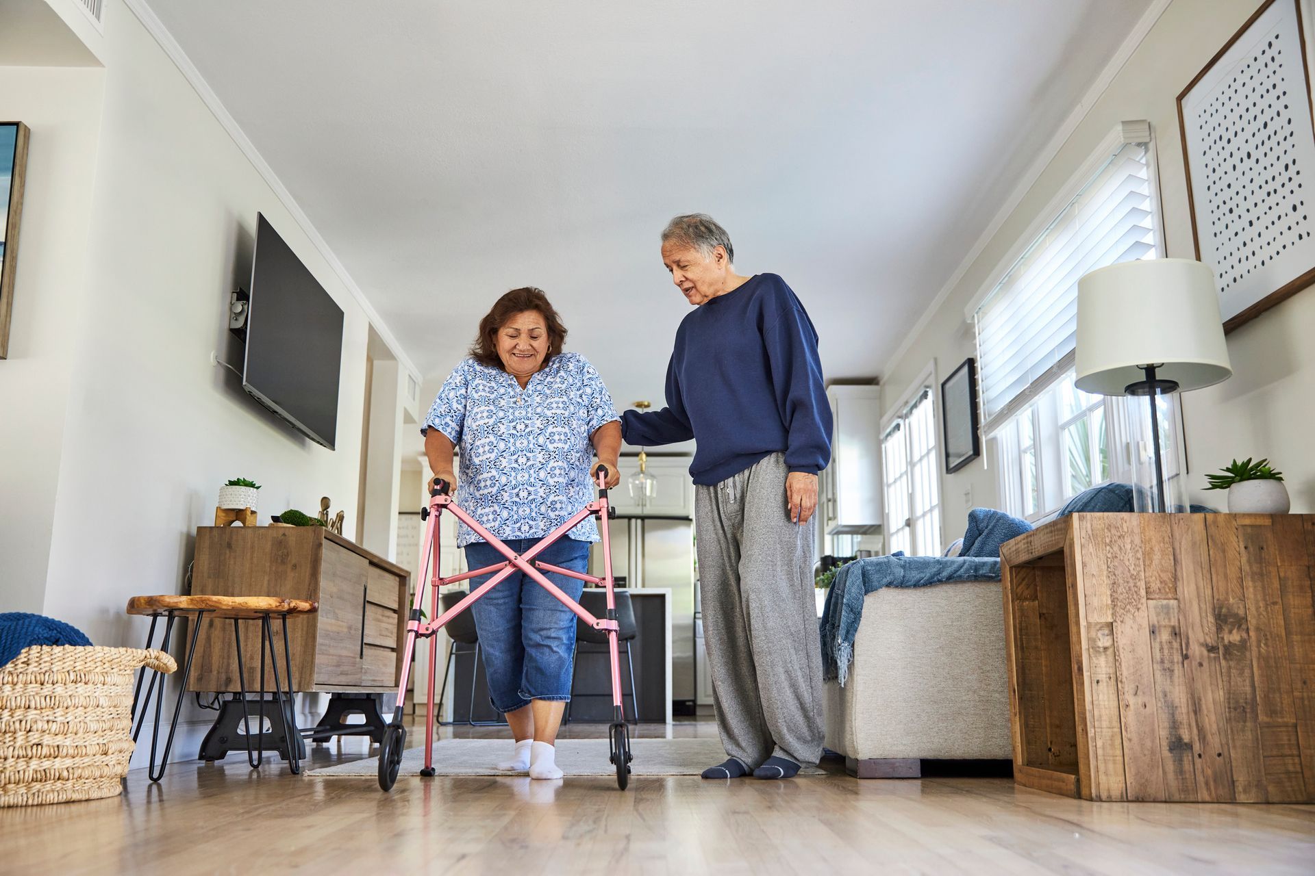 Senior woman walking with aid of pink walker from trusted mobility assistance equipment supplier. Woman using a walker with assistance from a man in a living room, smiling.