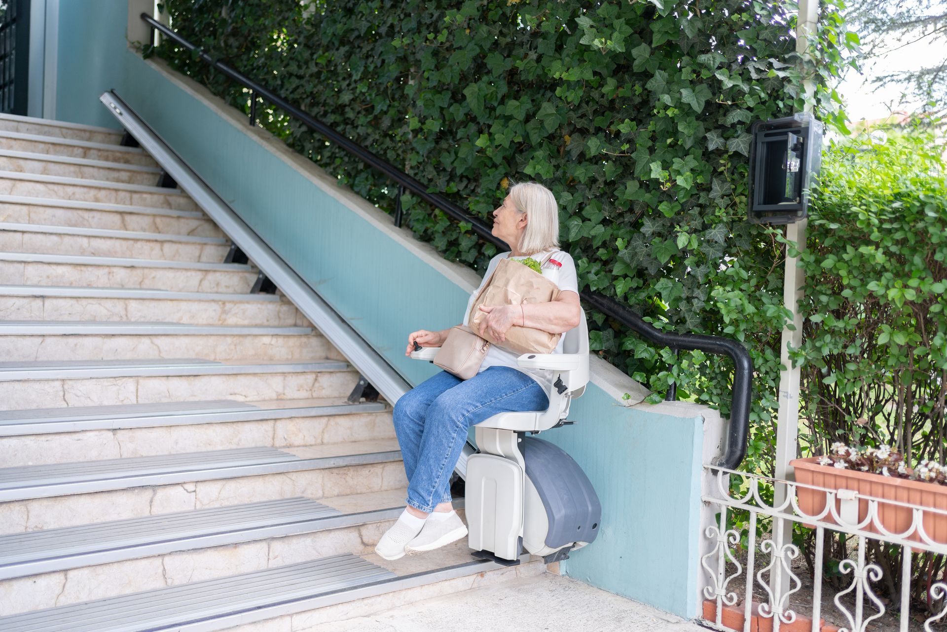 A senior woman uses an automatic stair lift, placed in a building with stairs at the entrance.