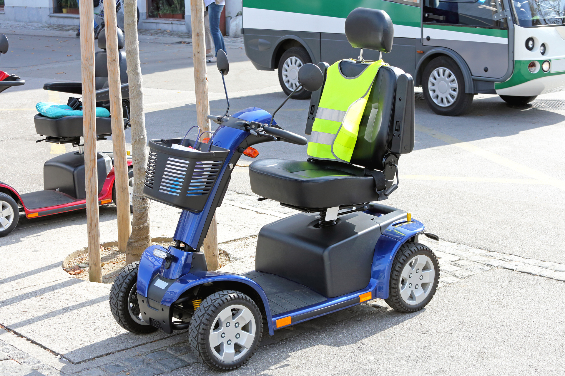 Blue mobility scooter with a yellow safety vest, parked outside near a bus.