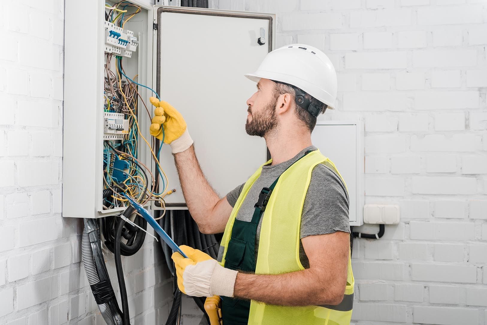 Technician in hard hat inspecting wiring inside an open electrical panel in a white room