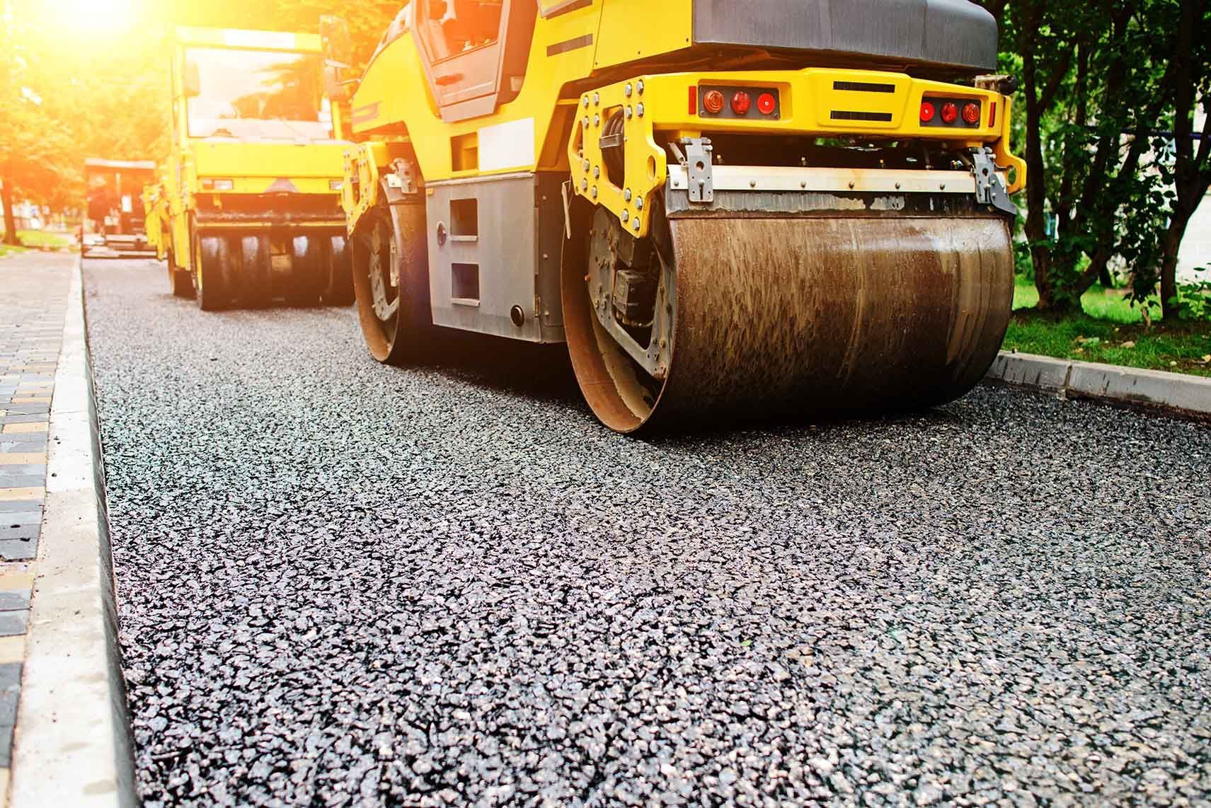 A road roller compresses fresh asphalt as paving work continues on a sunny day.