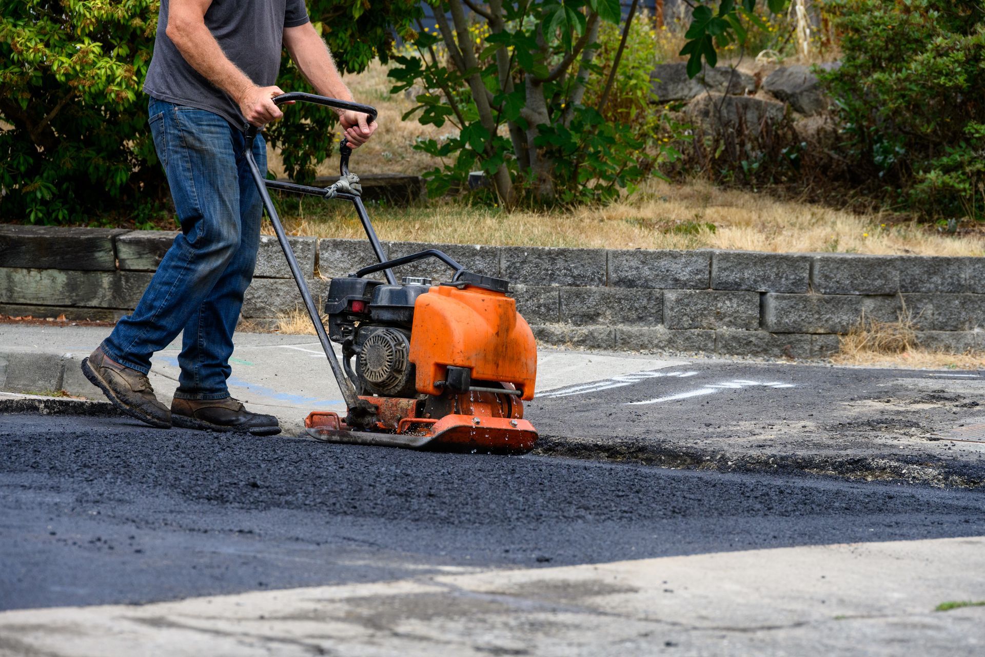 An asphalt driveway contractor works an orange plate compactor over fresh asphalt by a stone wall.