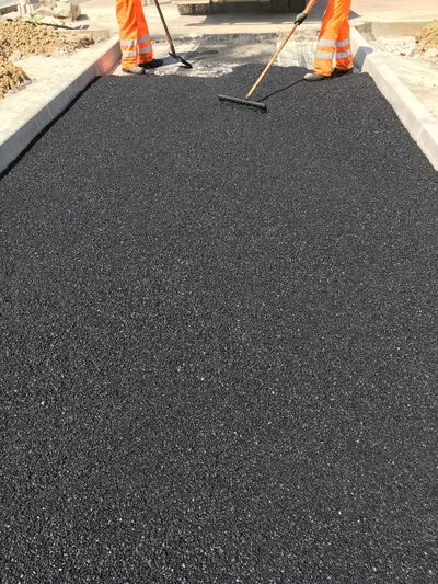 Two workers in high-visibility orange pants use long-handled rakes to spread fresh black asphalt on a narrow path.