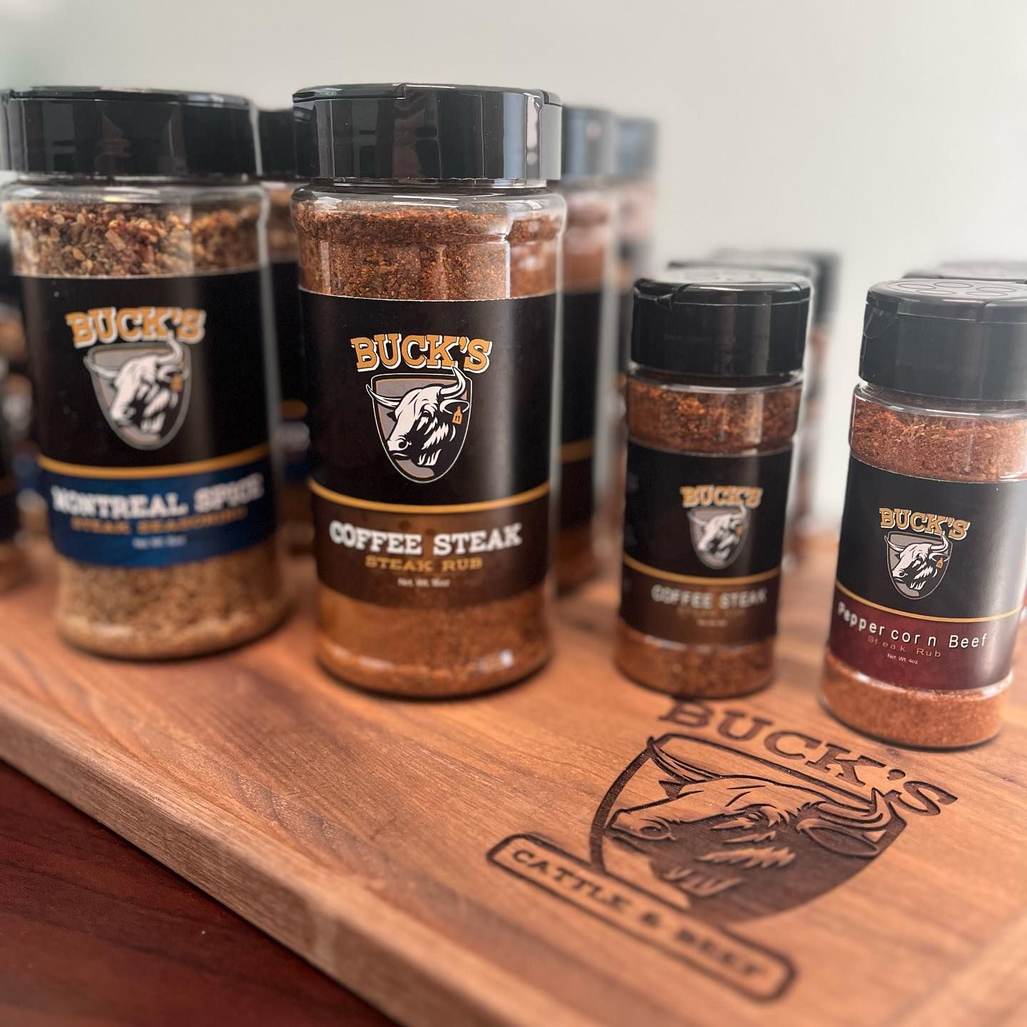 Spice jars on a wooden tray, including Buck’s coffee steak seasoning, with black lids and labels.