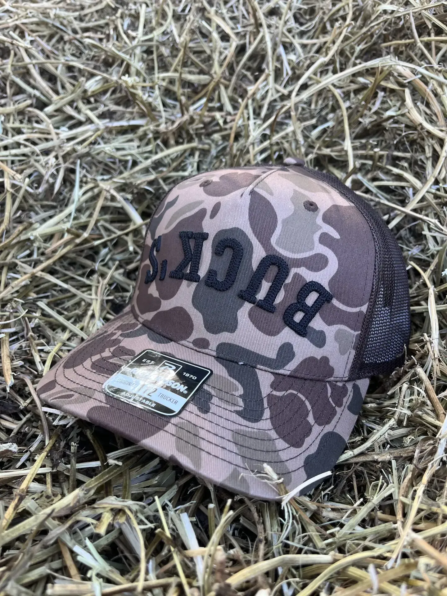 Camo baseball cap with bold black lettering resting on dry grass outdoors