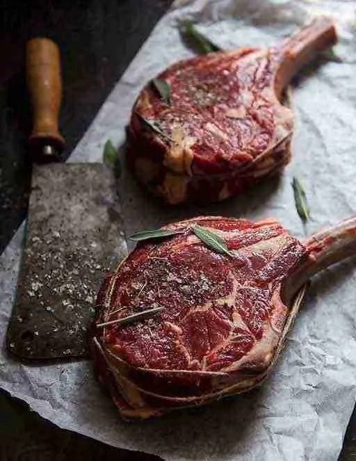Two raw ribeye steaks with marbling on parchment beside a meat cleaver