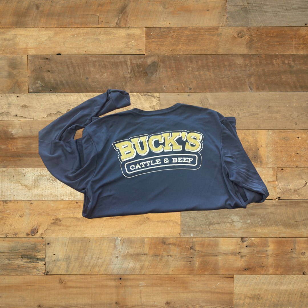 Folded navy T-shirt with yellow “BUCKS” and white “CATTLE & BEET” logo on a wooden floor