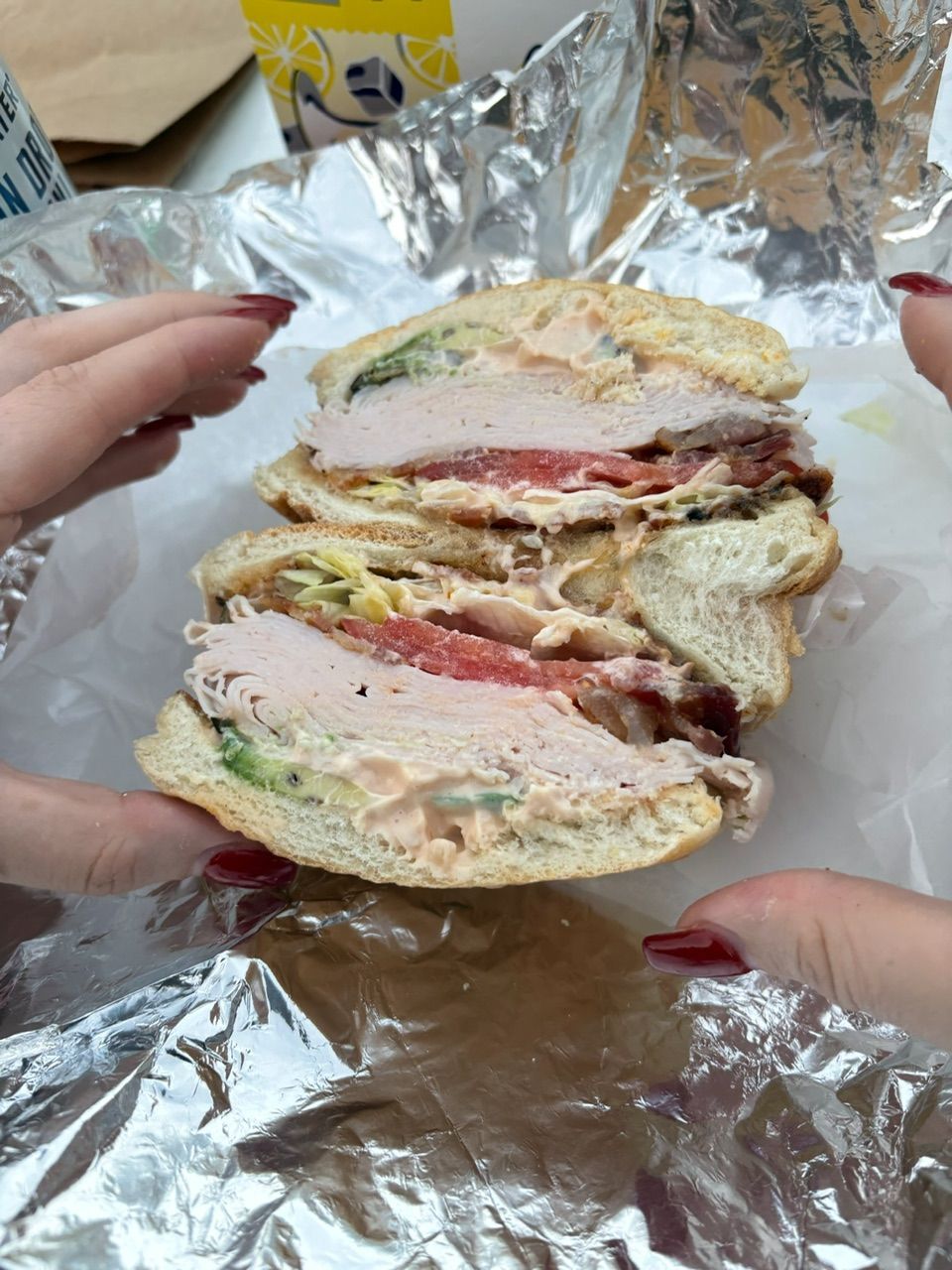 Hands holding a cut sandwich, filled with meat, tomato, and lettuce, wrapped in foil.
