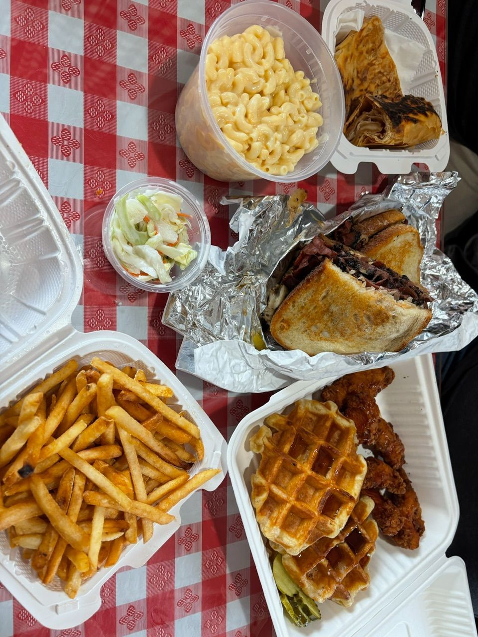 Overhead view of takeout meals on a red and white checkered table: fries, mac and cheese, sandwich, chicken, waffles, and coleslaw.