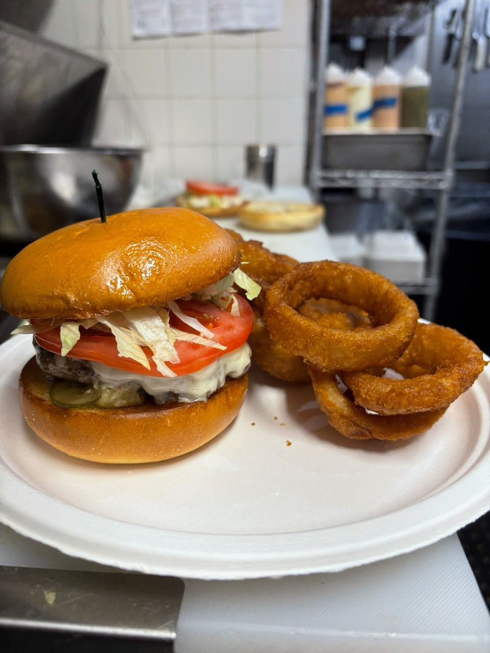 Hamburger on a bun with lettuce, tomato, and onion rings on a white plate. Background includes a stainless steel counter.