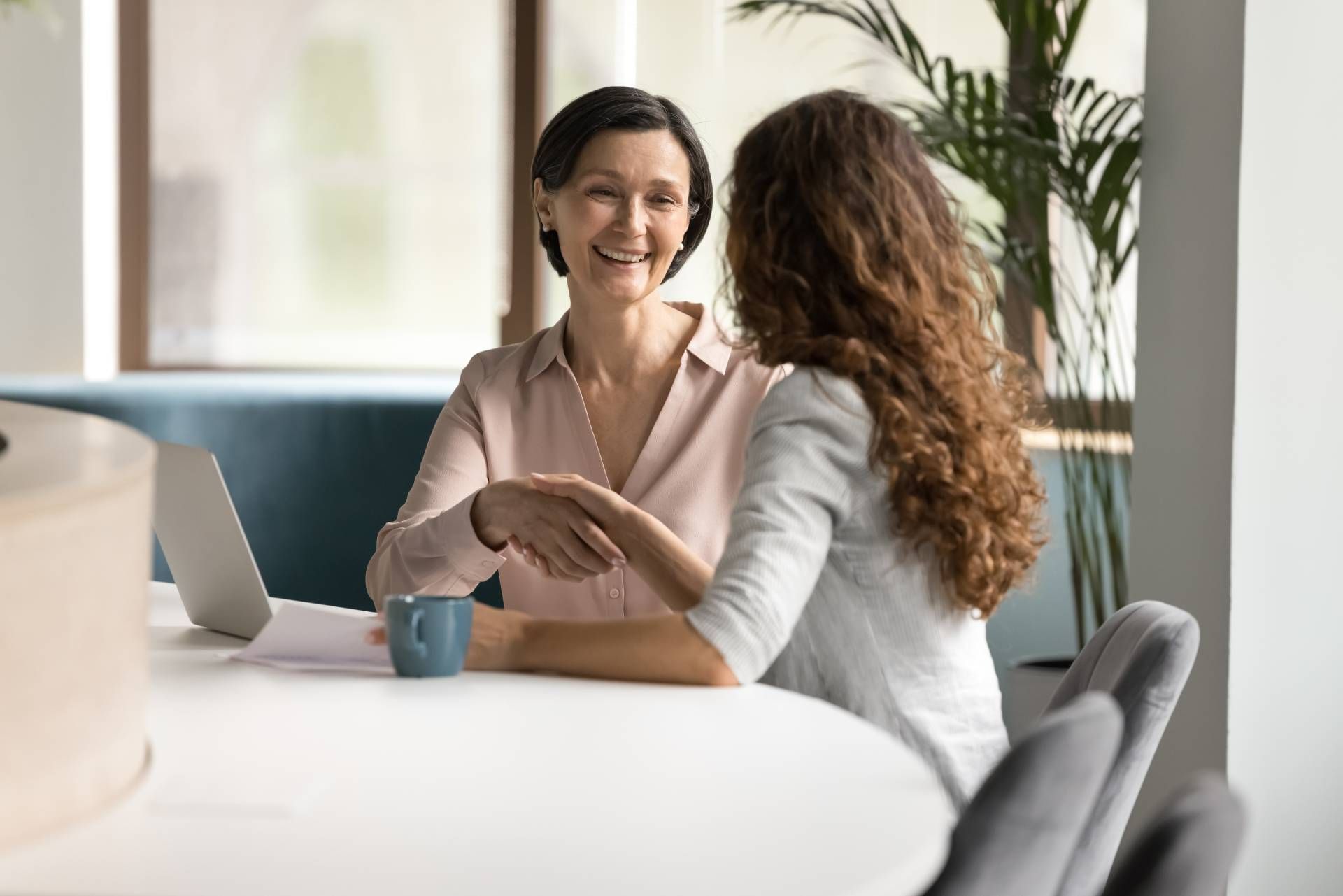 Two people talking at a table in a bright office, one smiling with a laptop nearby
