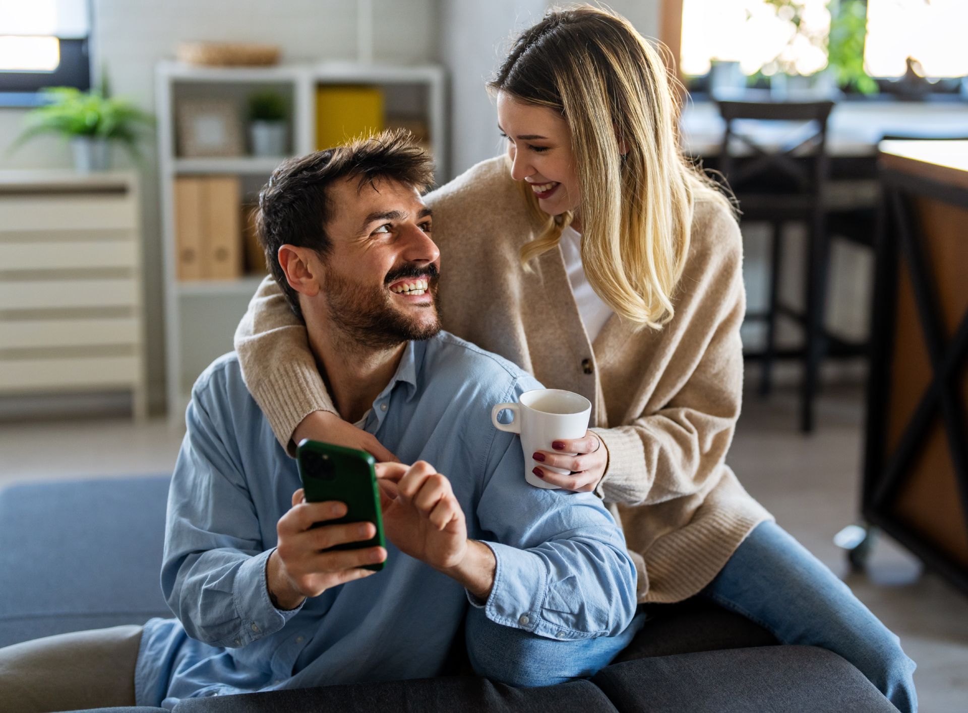Smiling couple on a couch, one holding a phone and the other a coffee cup in a cozy living room