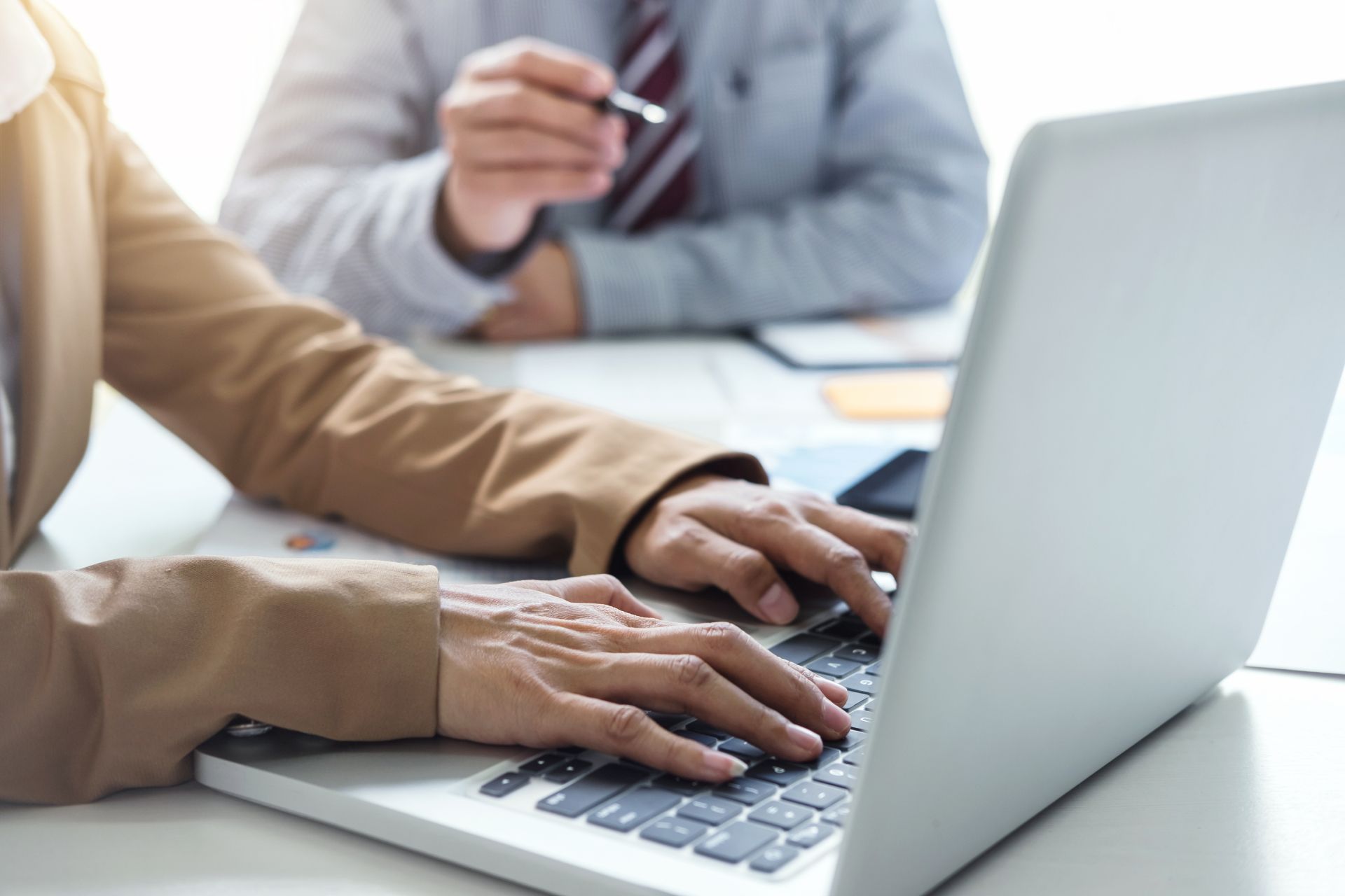 Hands typing on a laptop during a business meeting, with another person holding a pen in the background