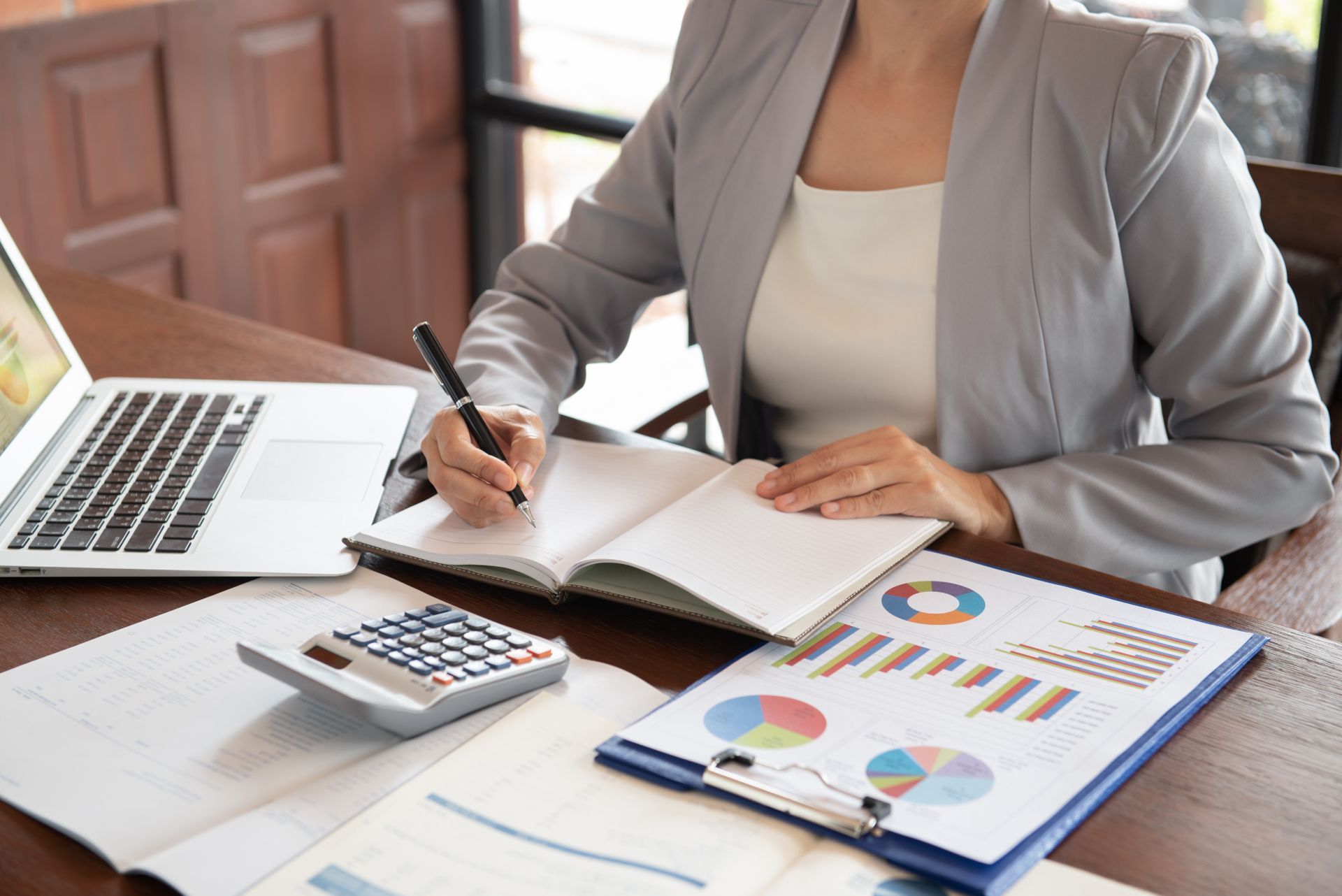 Person writing in a notebook beside a laptop, calculator, and financial charts on a desk